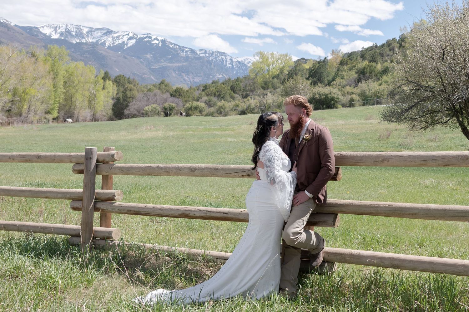 Bride and groom share romantic moment by rustic fence with snow-capped mountains backdrop.