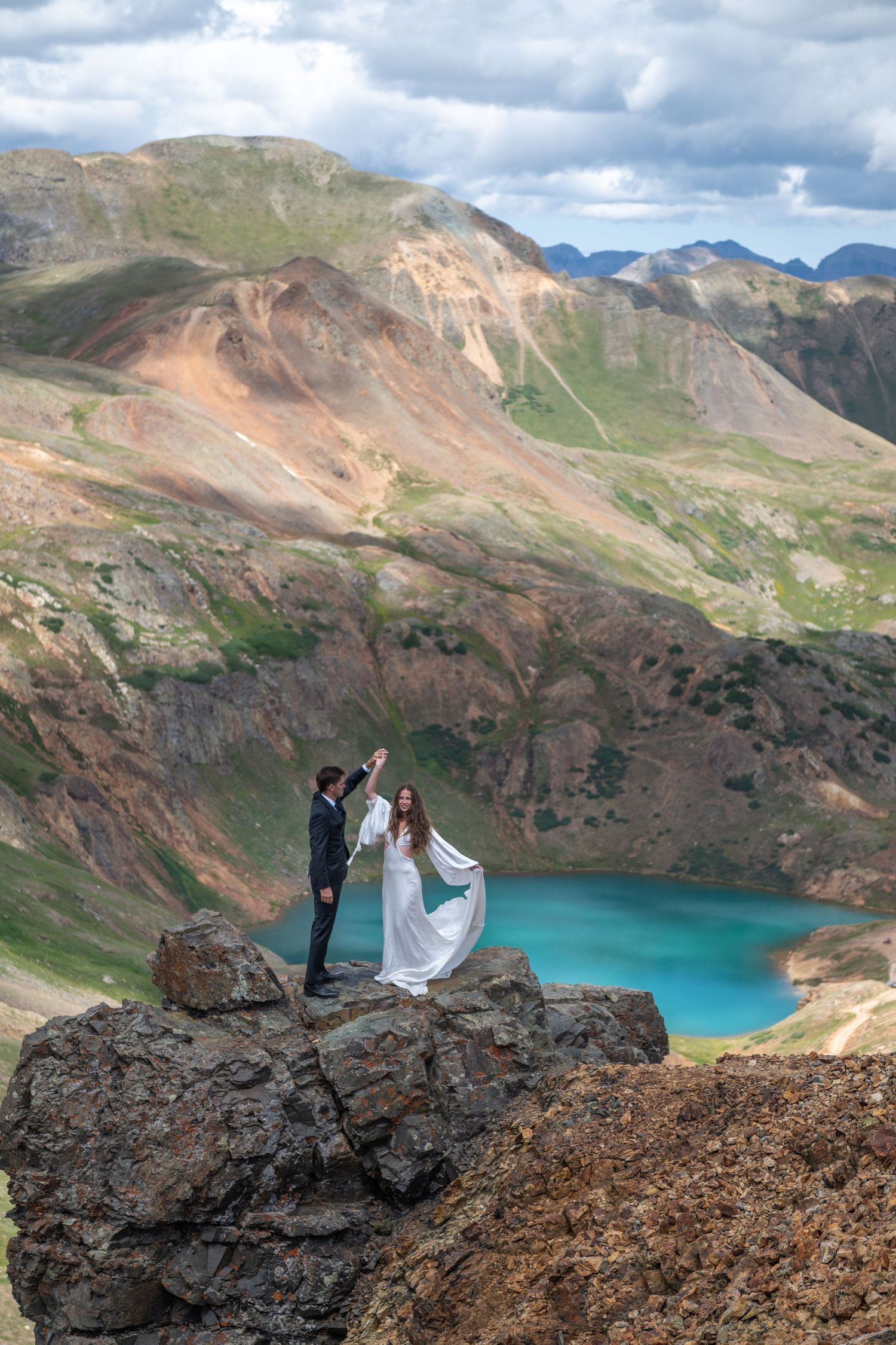 Couple dancing on rocky mountain peak with turquoise alpine lake and colorful mountains in background during outdoor wedding.