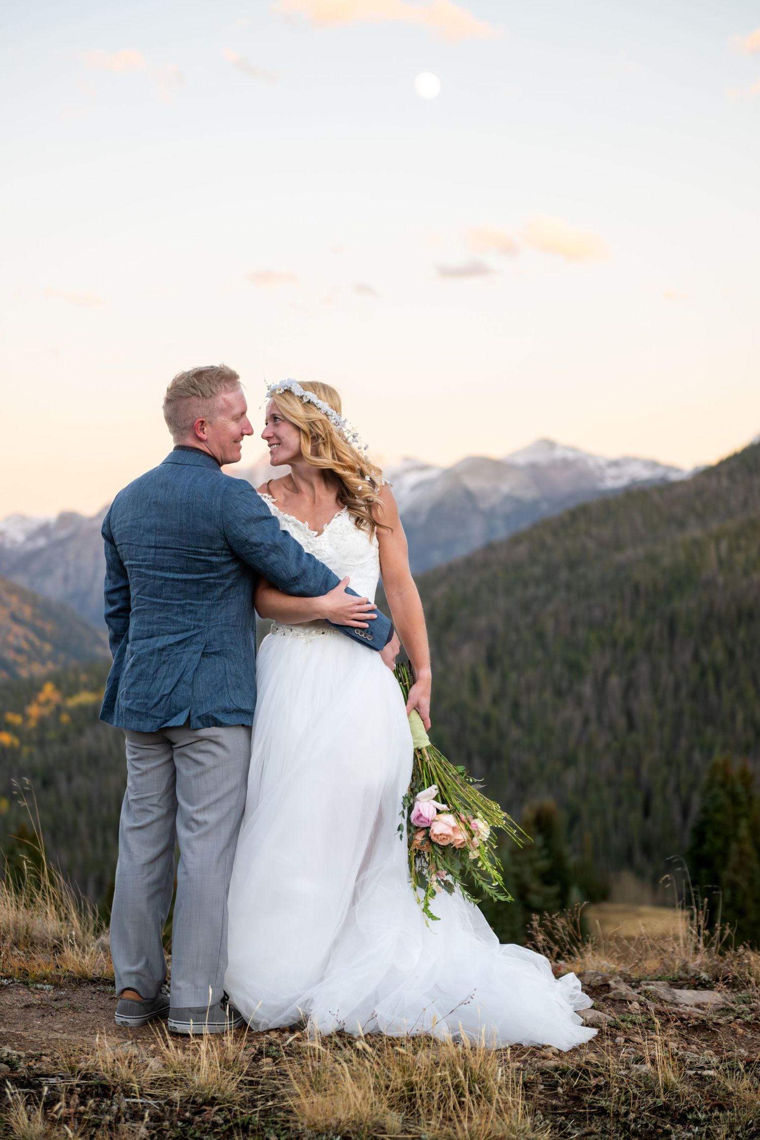 A couple in wedding attire embrace while standing on a mountain overlook at sunset with snow-capped mountain peaks in the distance.