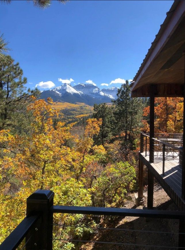 Snow-capped mountain peaks rise above autumn trees with yellow foliage seen through a rustic Airbnb cabin porch frame near Ouray.
