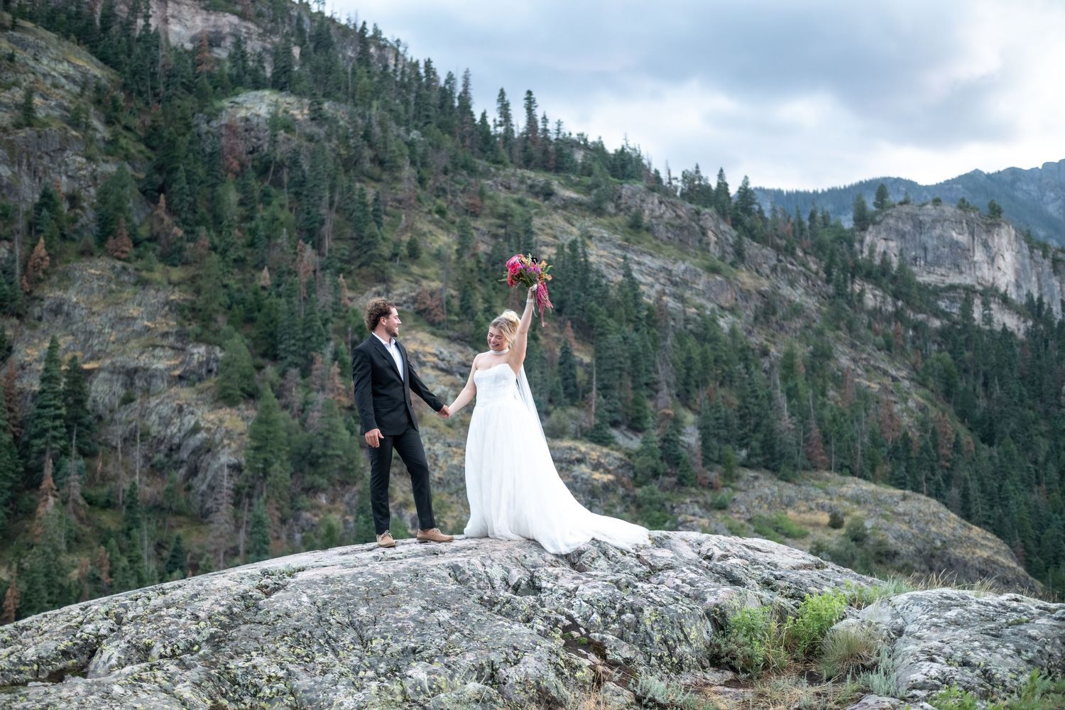 Bride and groom stand on rocky mountain summit holding bouquet aloft with evergreen forest backdrop.