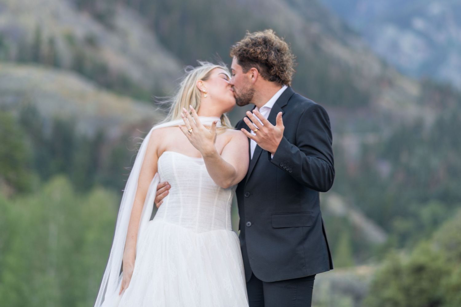 Bride and groom sharing a kiss outdoors with mountain scenery, bride in white gown with veil, groom in dark suit.