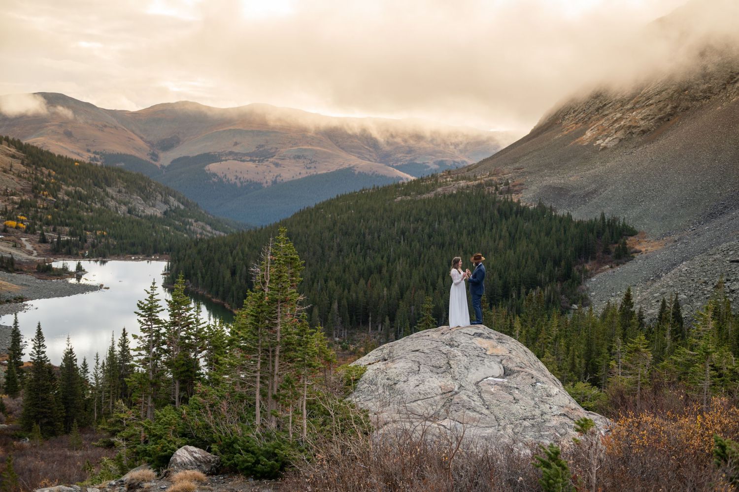 An eloping couple in wedding attire standing on a boulder with mountain lake and forest views near Breckenridge