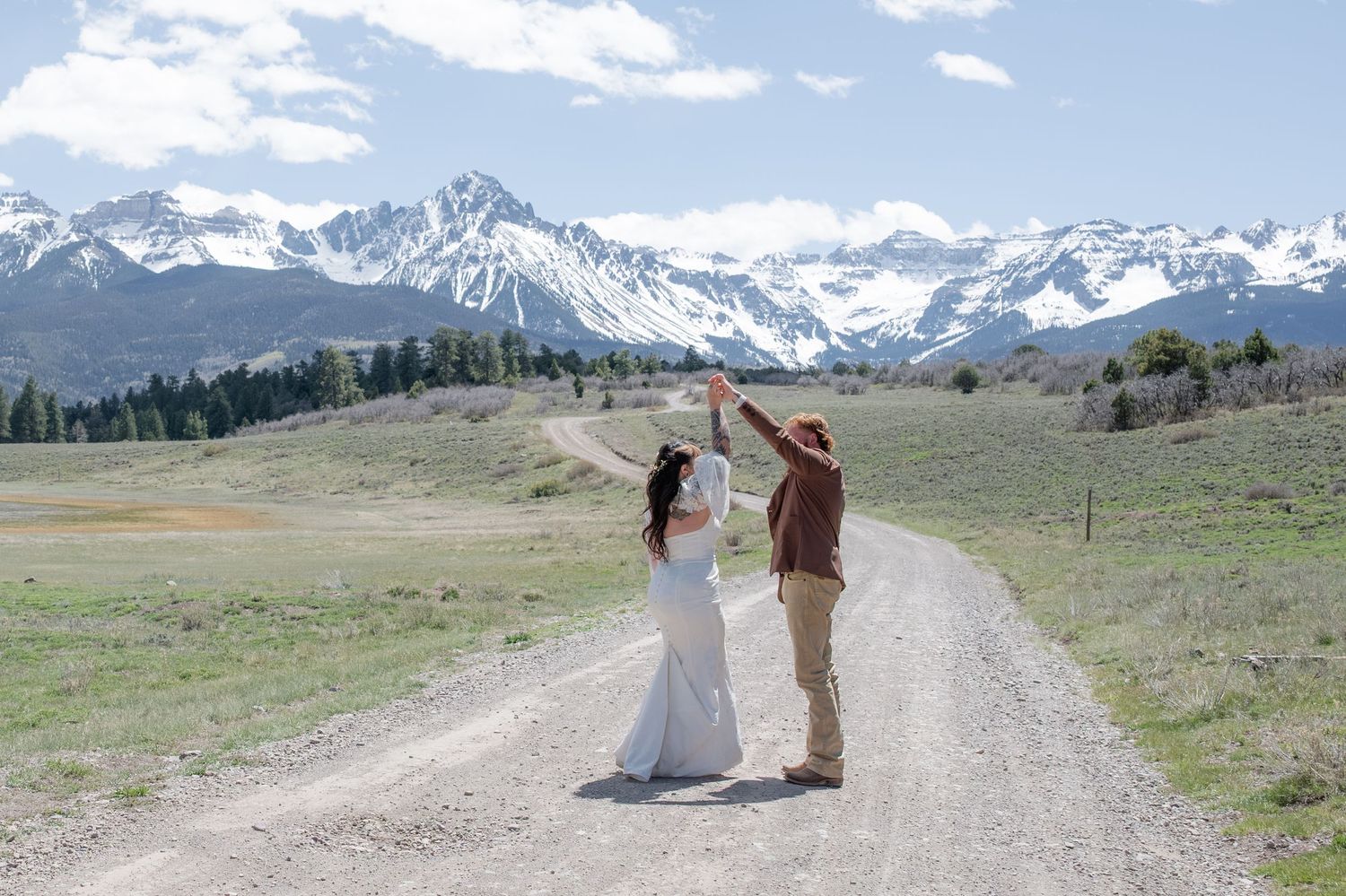 Bride and groom dancing on a mountain dirt road with snow-capped peaks and green meadows in the background.