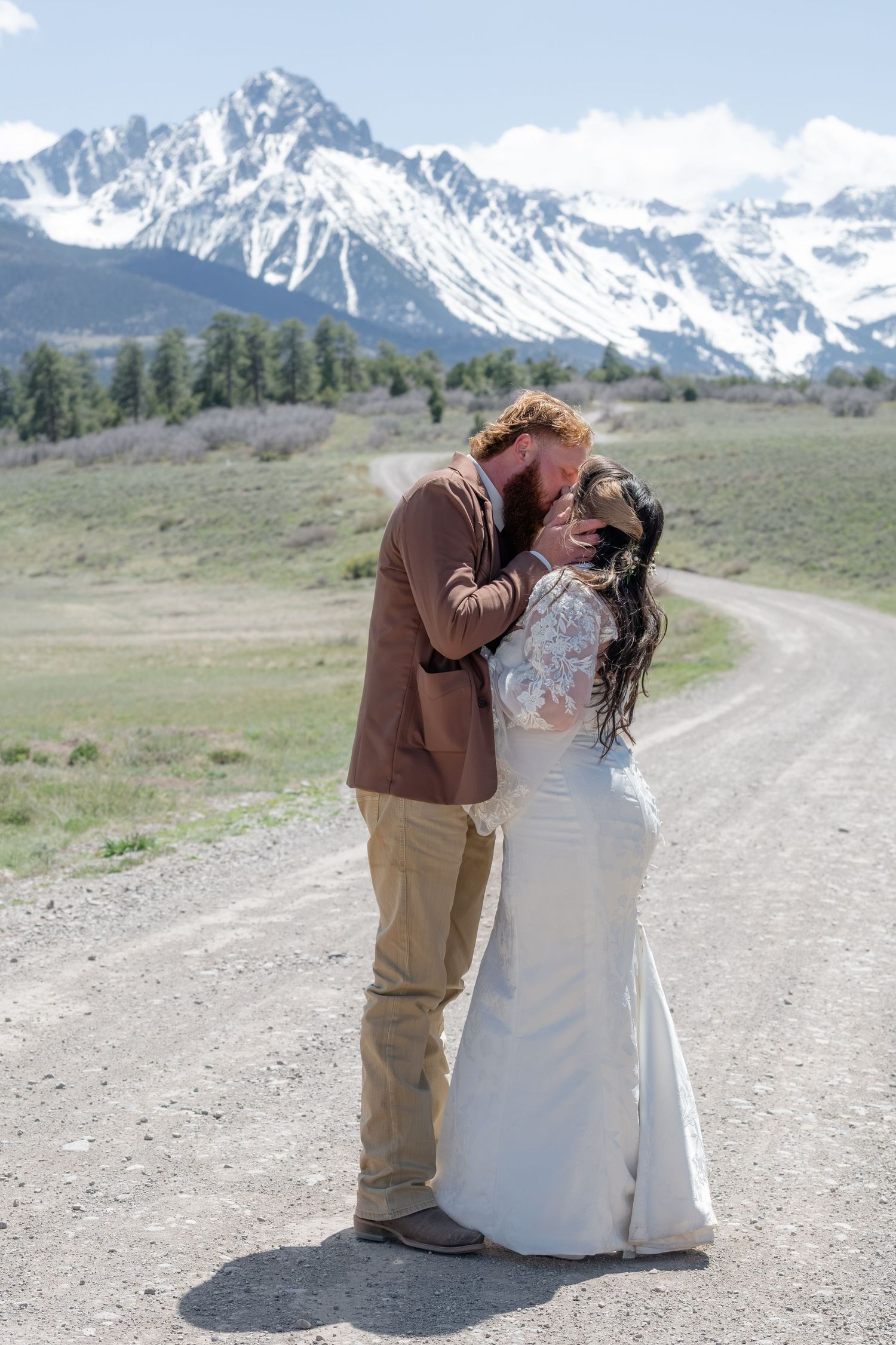 A groom kisses his bride in the middle of a dirt road with the San Juan Mountains in the background. Snow is on the peaks of the mountains.