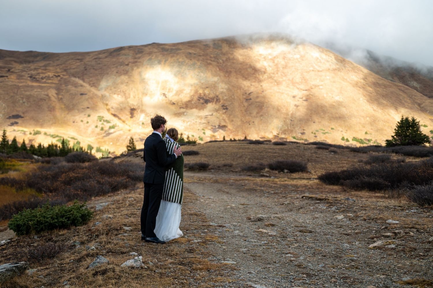 A bride and groom stand in an embrace after their Breckenridge elopement. Her back is to his chest. She is wrapped in a striped blanket.