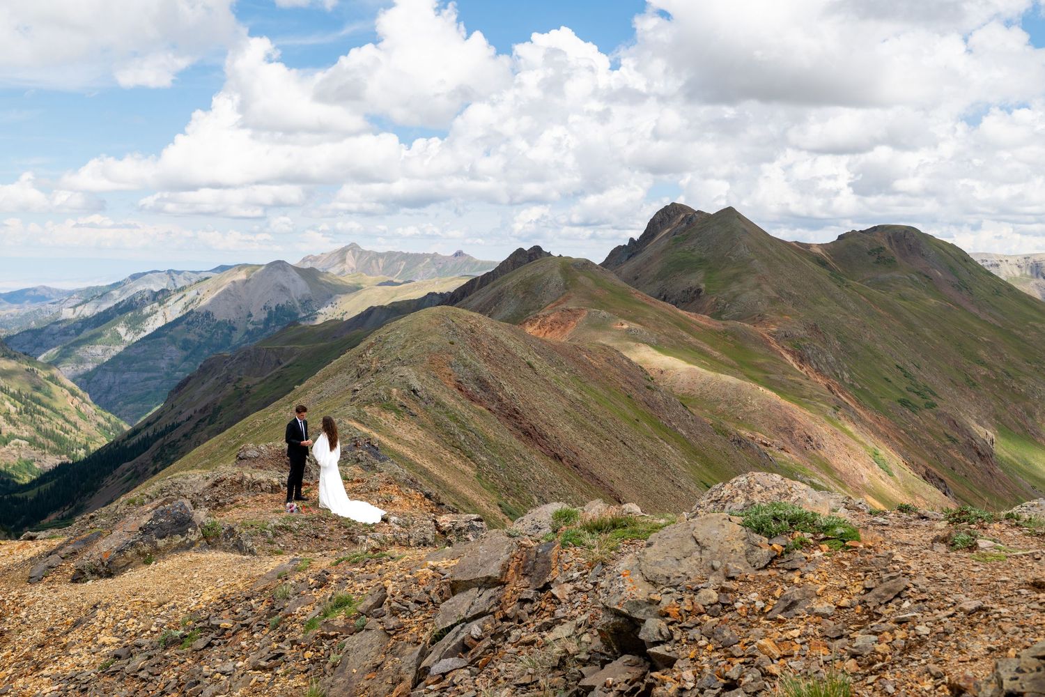 Bride and groom stand atop a rocky mountain peak surrounded by stunning green and red mountain ranges under a cloudy sky.