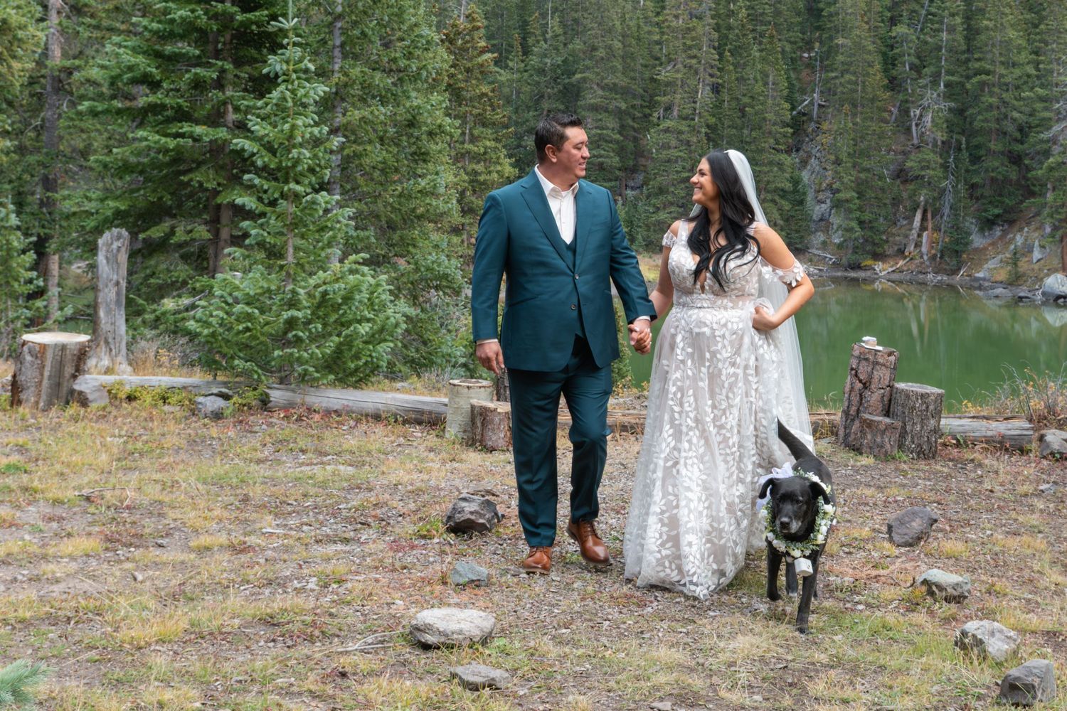 Bride and groom walk hand-in-hand after their elopement ceremony with their black dog near a mountain lake surrounded by pine trees.