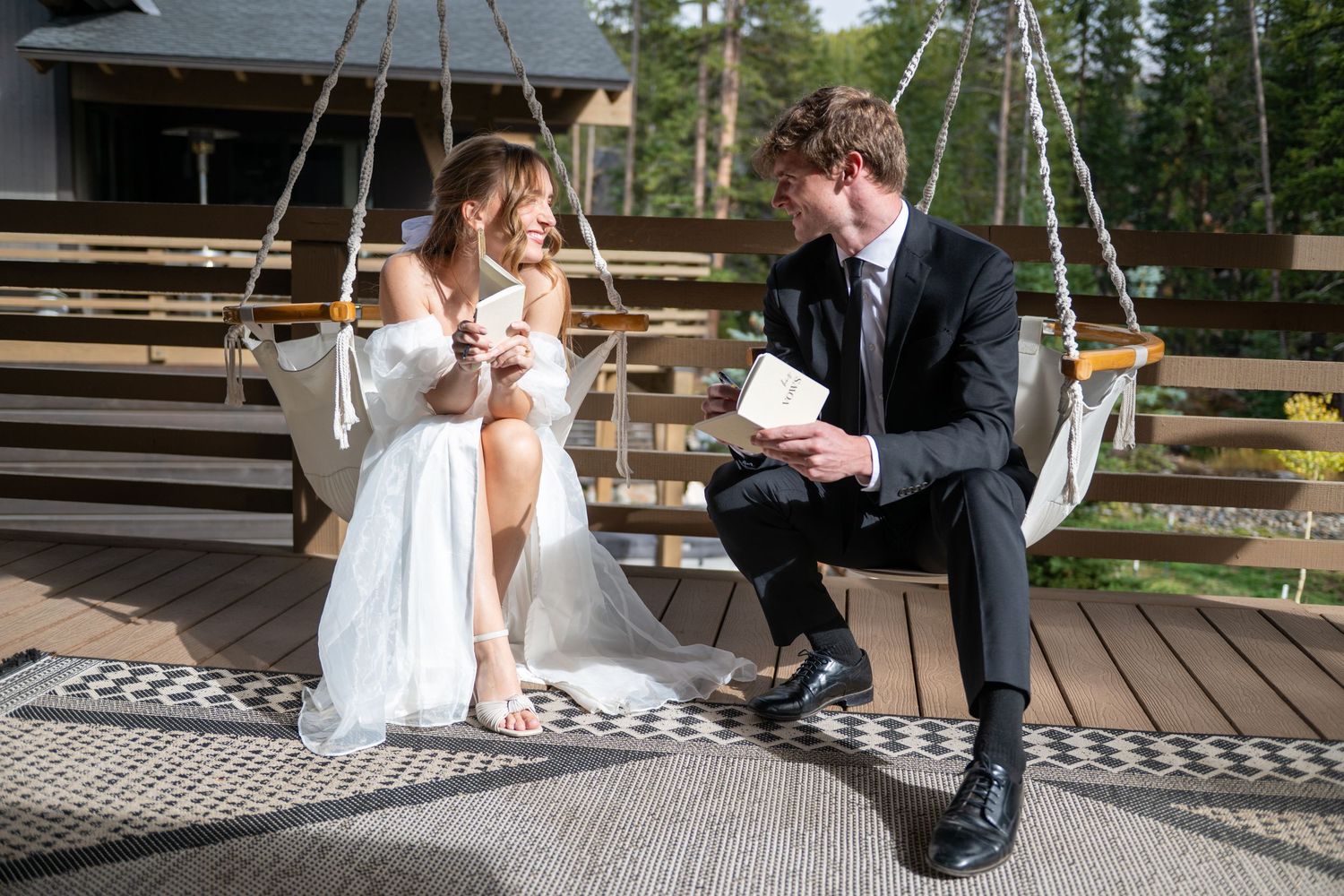 An eloping couple is dressed in wedding attire sitting in hammock swings on the balcony of a mountain cabin. They are smiling at one another