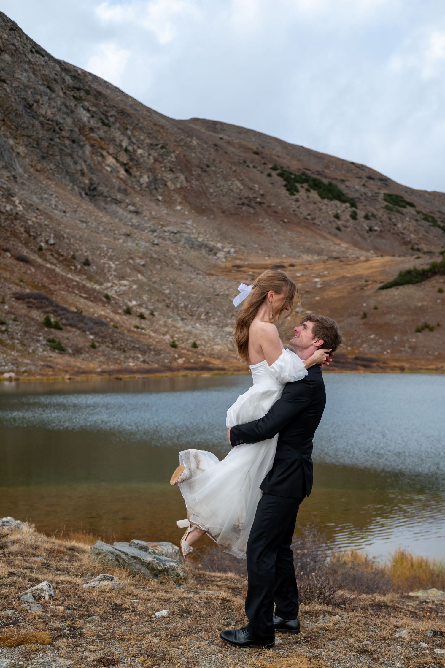 A groom lifting his bride in a white dress by a high mountain lake during outdoor elopement wedding. She has a white bow in her hair.