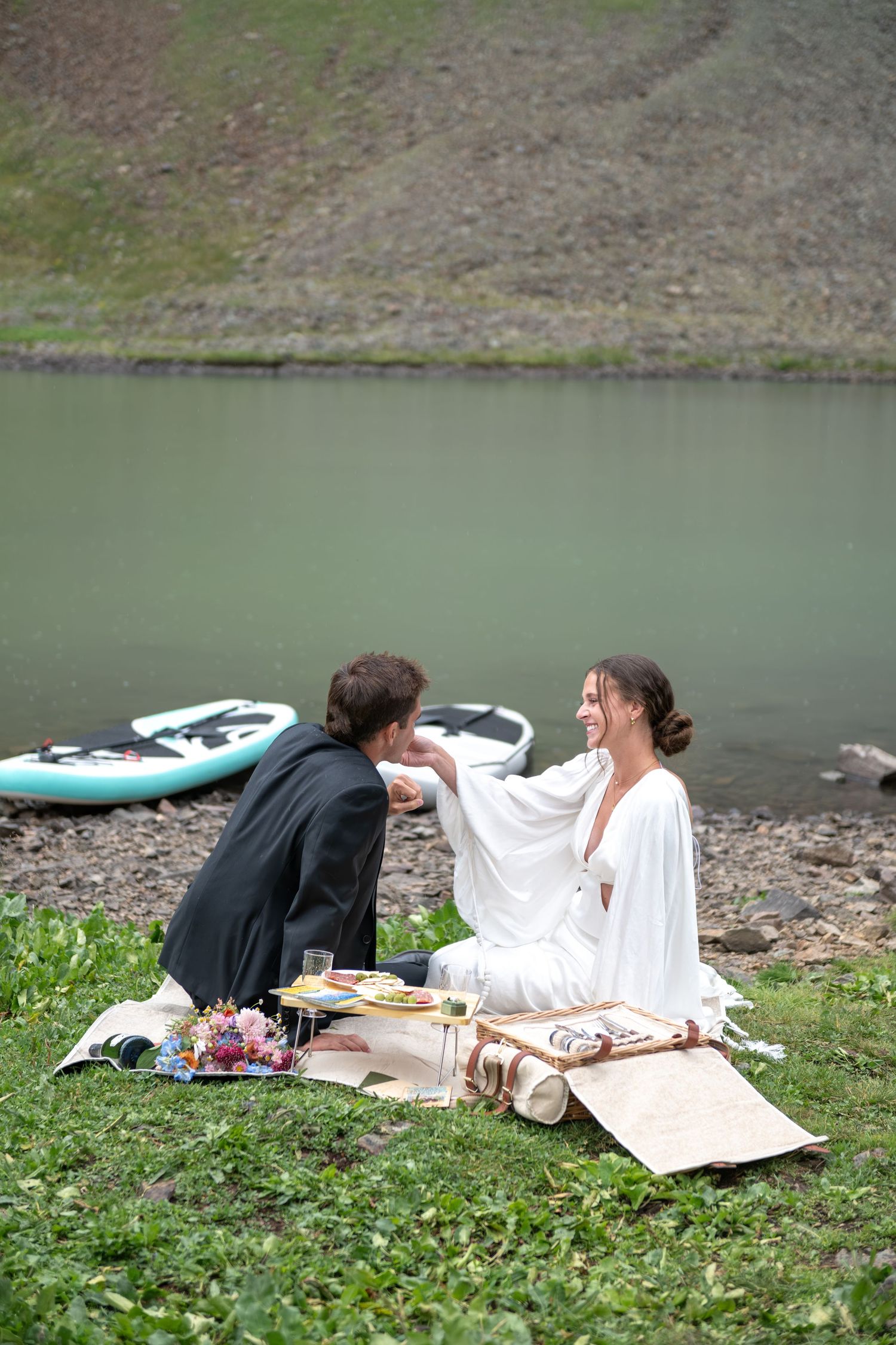 Bride and groom sharing a romantic lakeside picnic on grass, with paddleboards visible near the water.