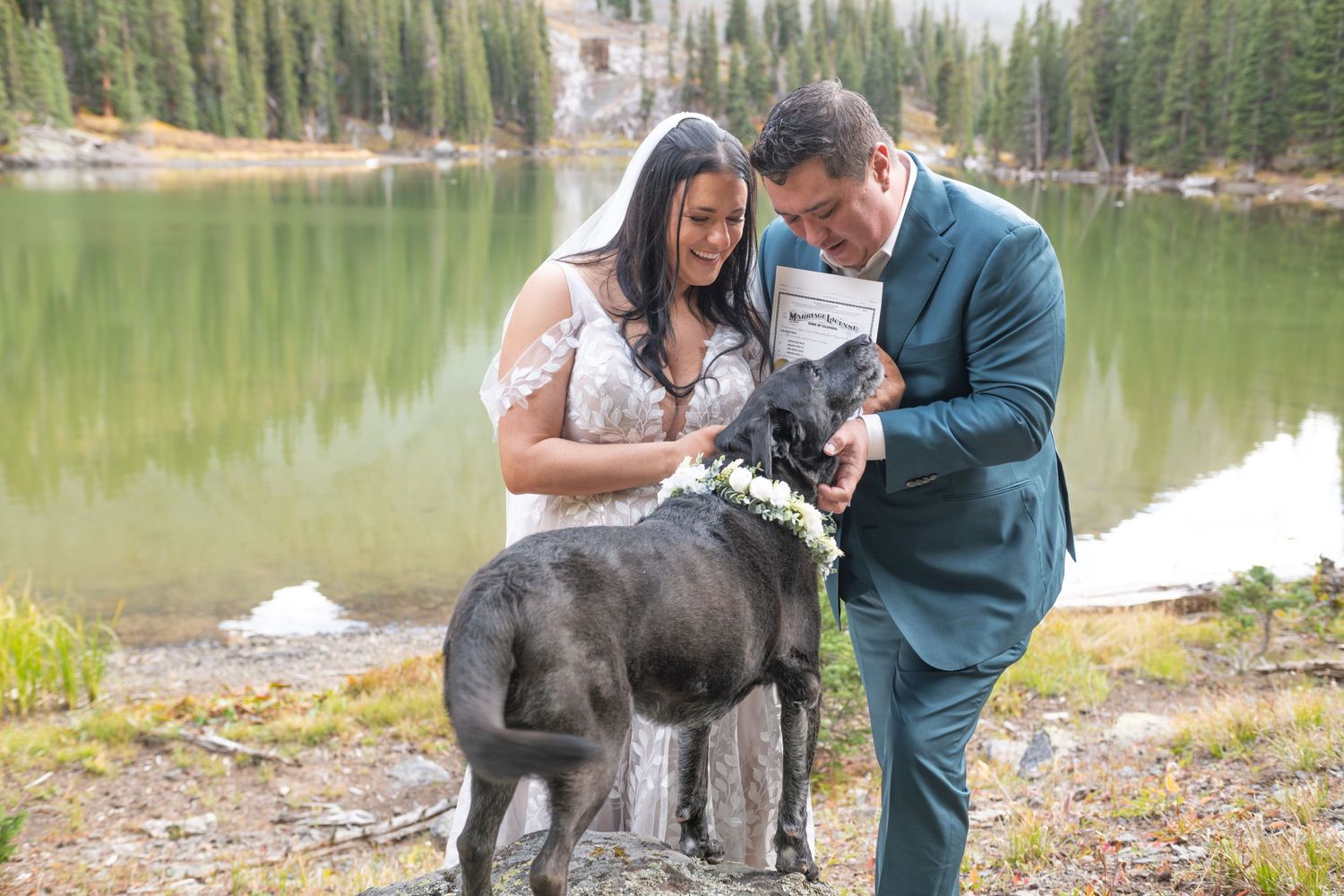 A bride and groom share a happy moment with their dog after their vows and the dog has signed the marriage license beside an alpine lake.