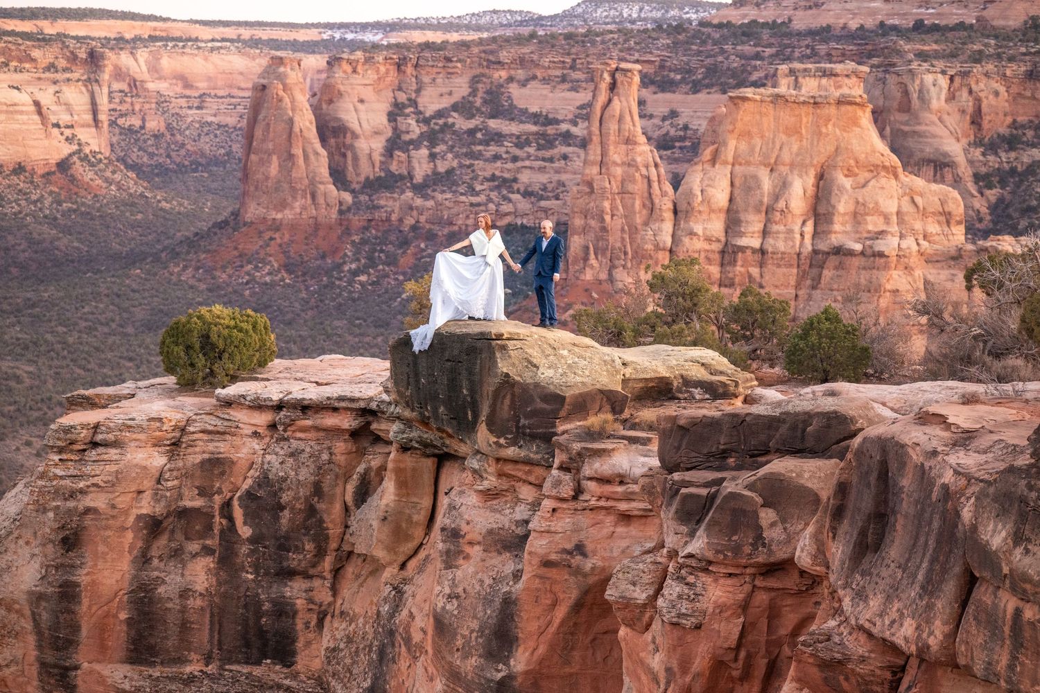 Aride and groom stand on a red rock cliff edge at Colorado National Monument with dramatic canyon views.