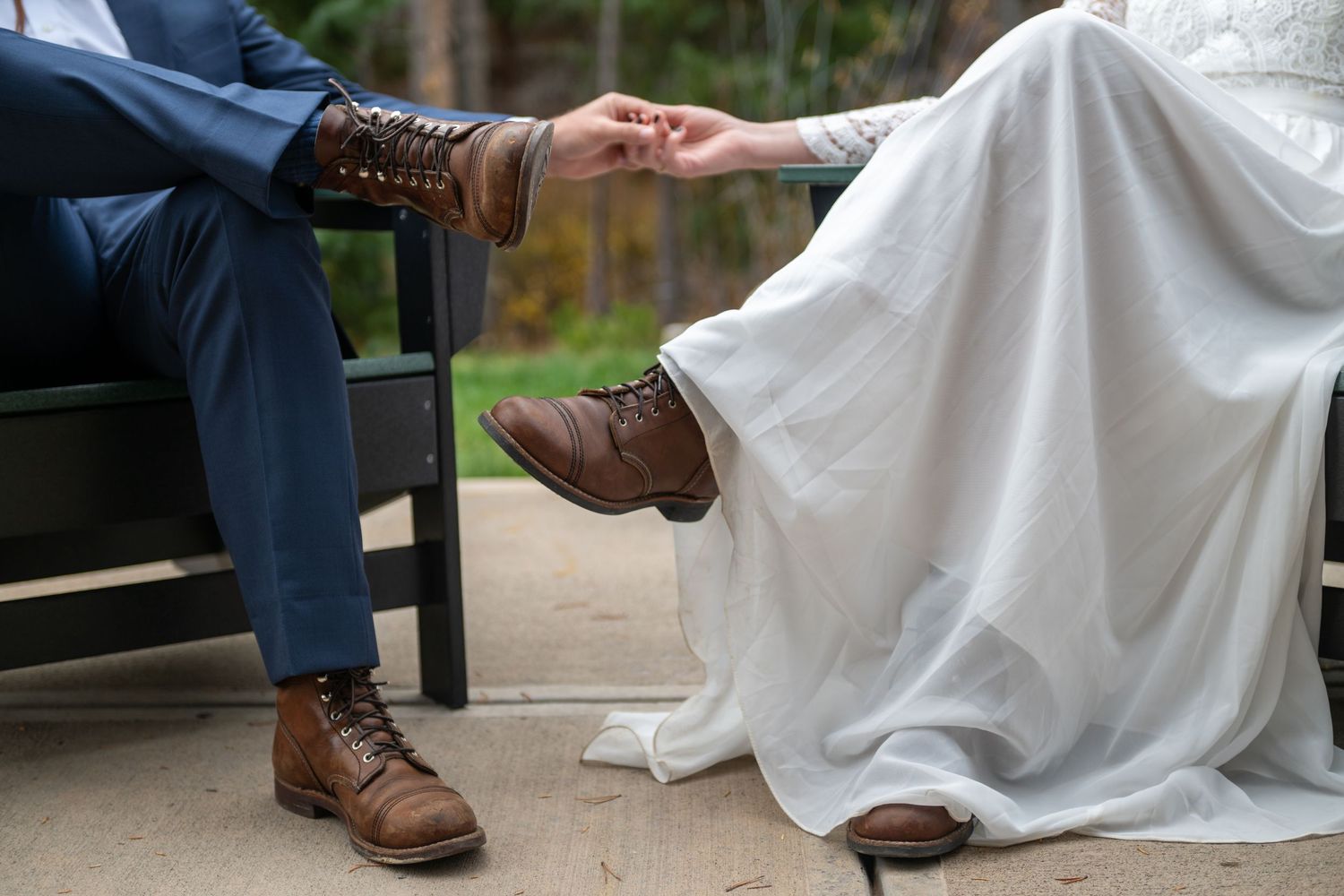 Bride in white gown and groom in blue suit holding hands, both wearing brown leather boots. they are sitting in green Adirondack chairs.