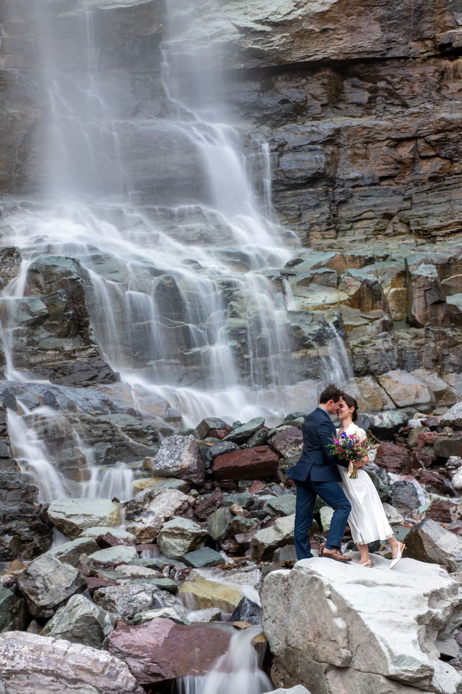 Bride and groom share a romantic kiss on rocky terrain in front of a stunning cascading waterfall near Ouray on their elopement day.