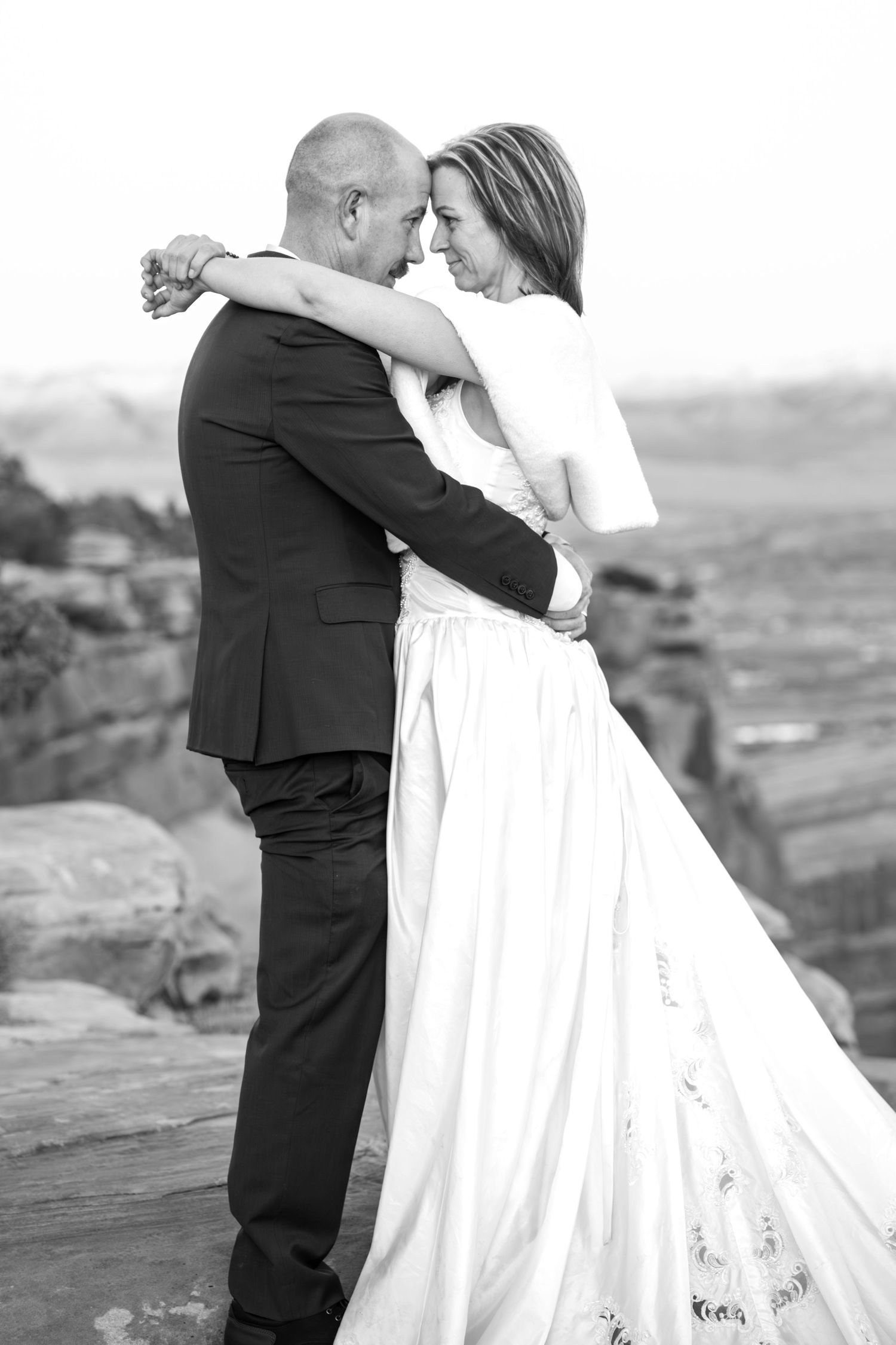 Black and white photo of a bride and groom embracing on the edge of a rock cliff. They are chest to chest with her arms around his neck.
