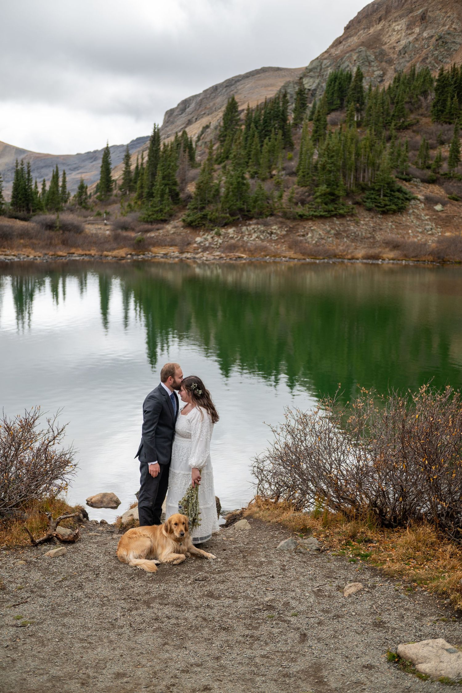 Couple in wedding attire sharing a kiss by a reflective mountain lake with their golden retriever dog nearby.