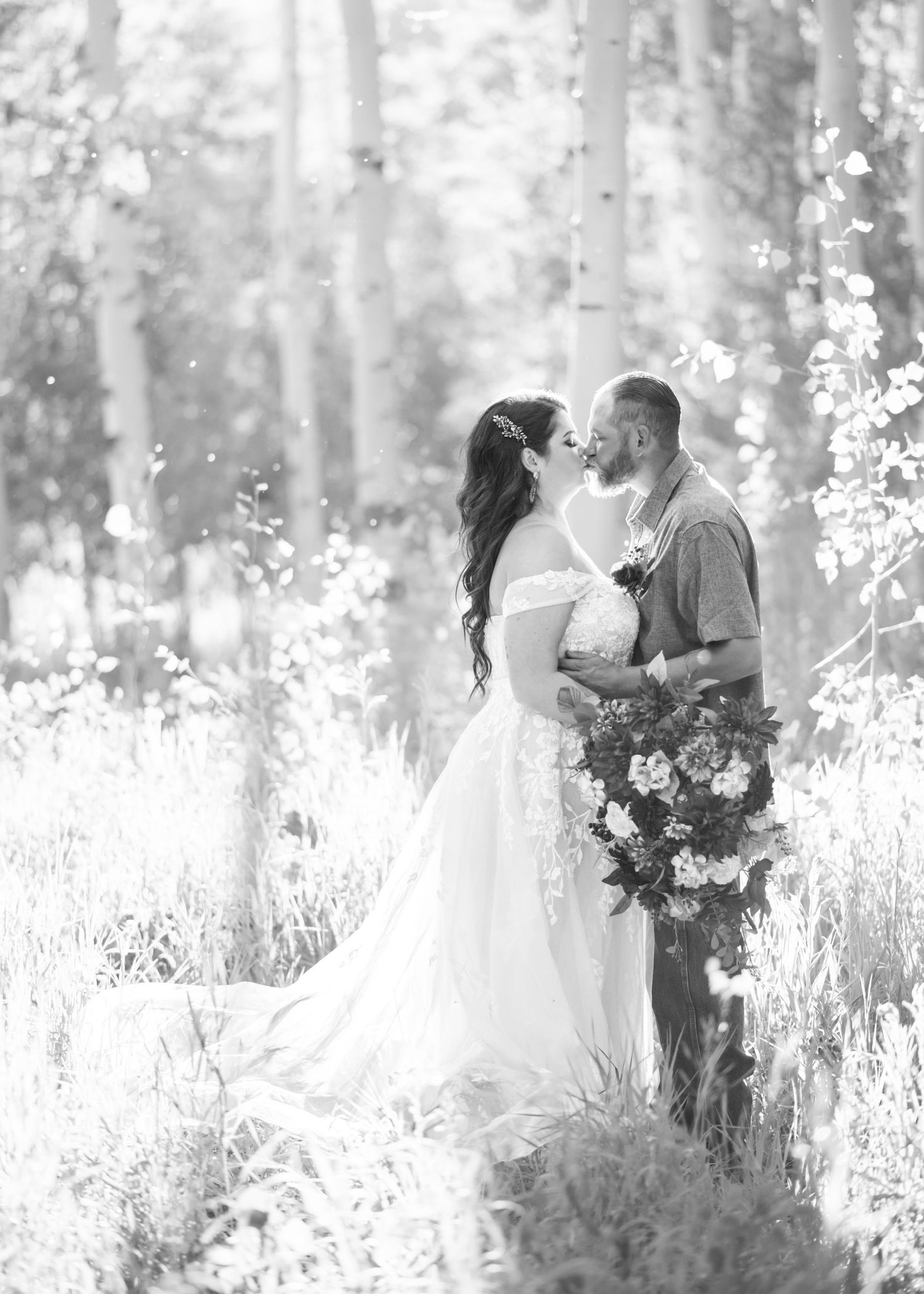 Black and white photo of a bride and groom sharing a kiss in a lush forest with aspens behind them. bride is holding a bouquet.