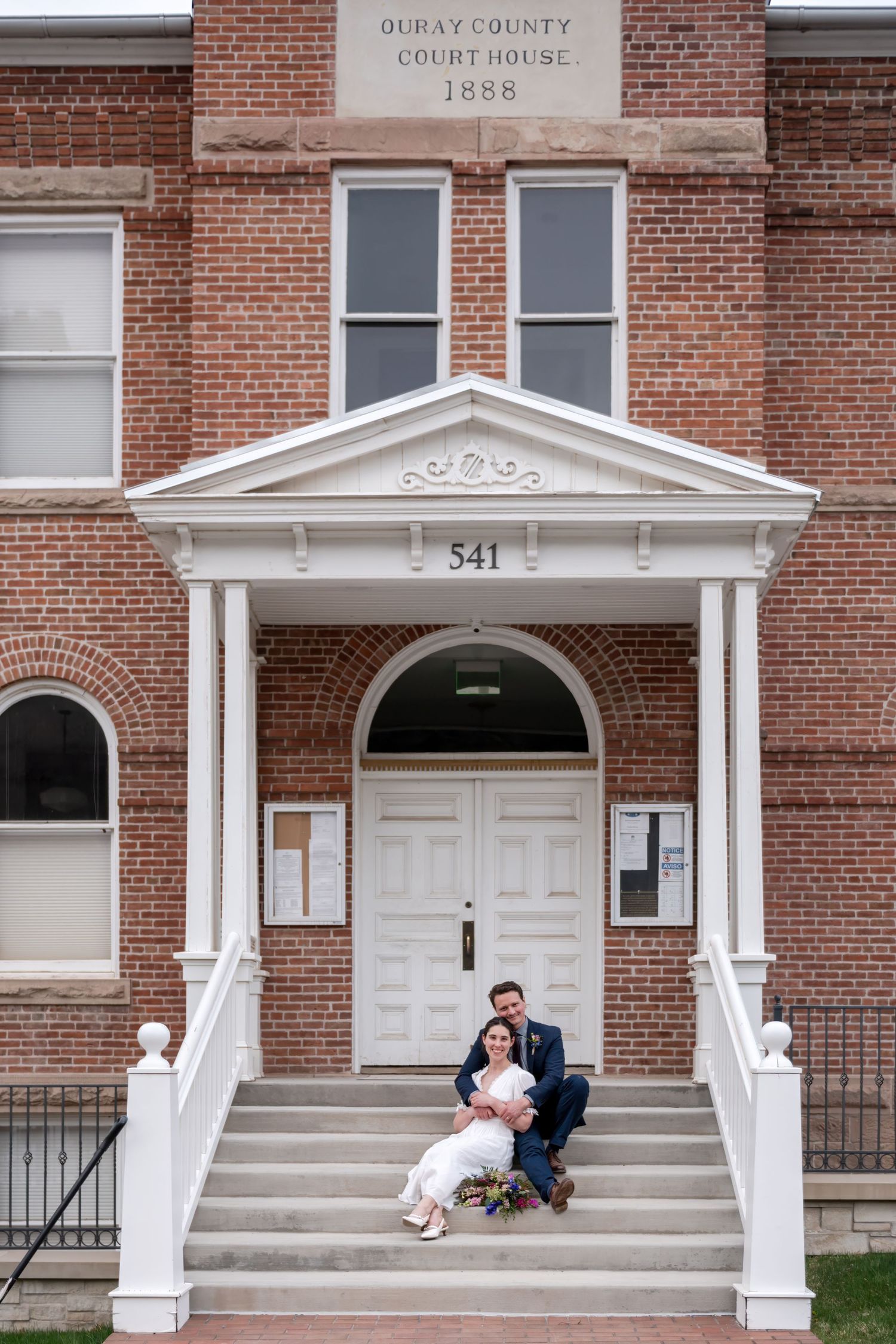 A bride and groom sit on the steps of the courthouse in Ouray Colorado where they said their vows. They are dressed in wedding attire.