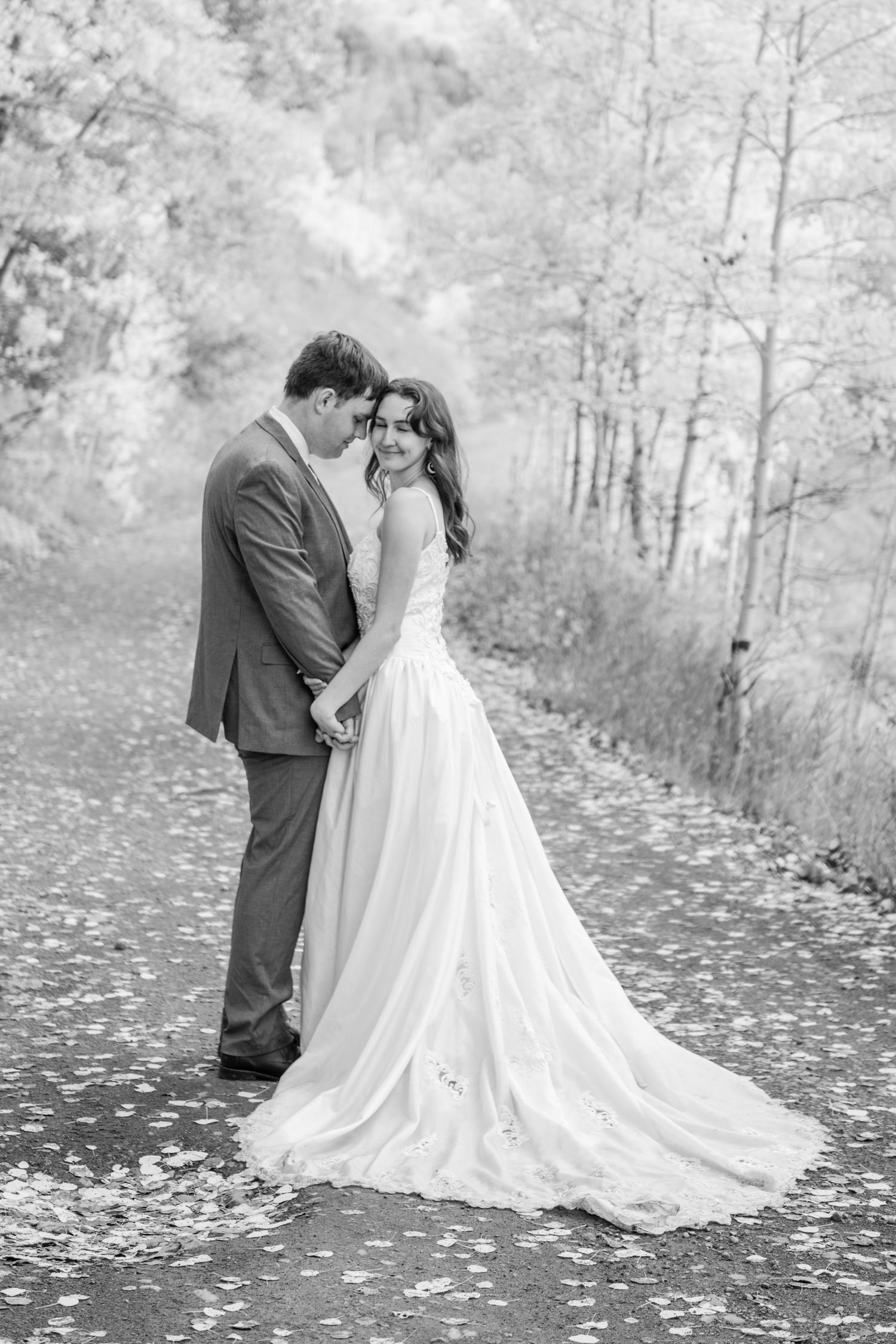 Black and white photo of a bride and groom embracing on a tree-lined path, the bride in a flowing gown looking back at the camera.