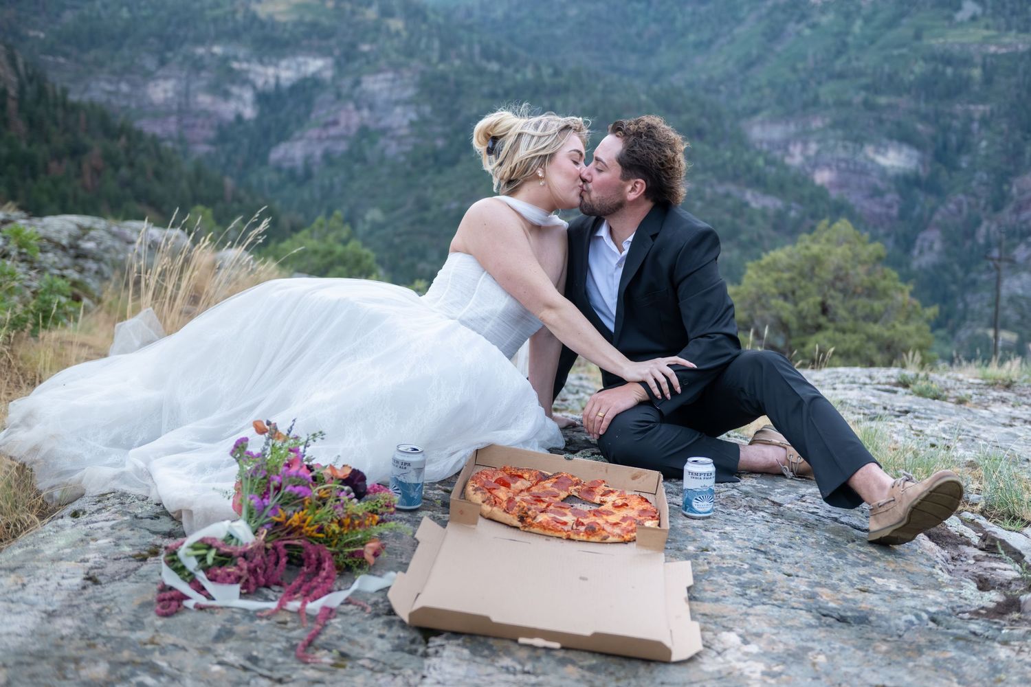 Newlywed couple kissing on a mountain cliff with pizza and flowers, celebrating their wedding with a scenic valley view.