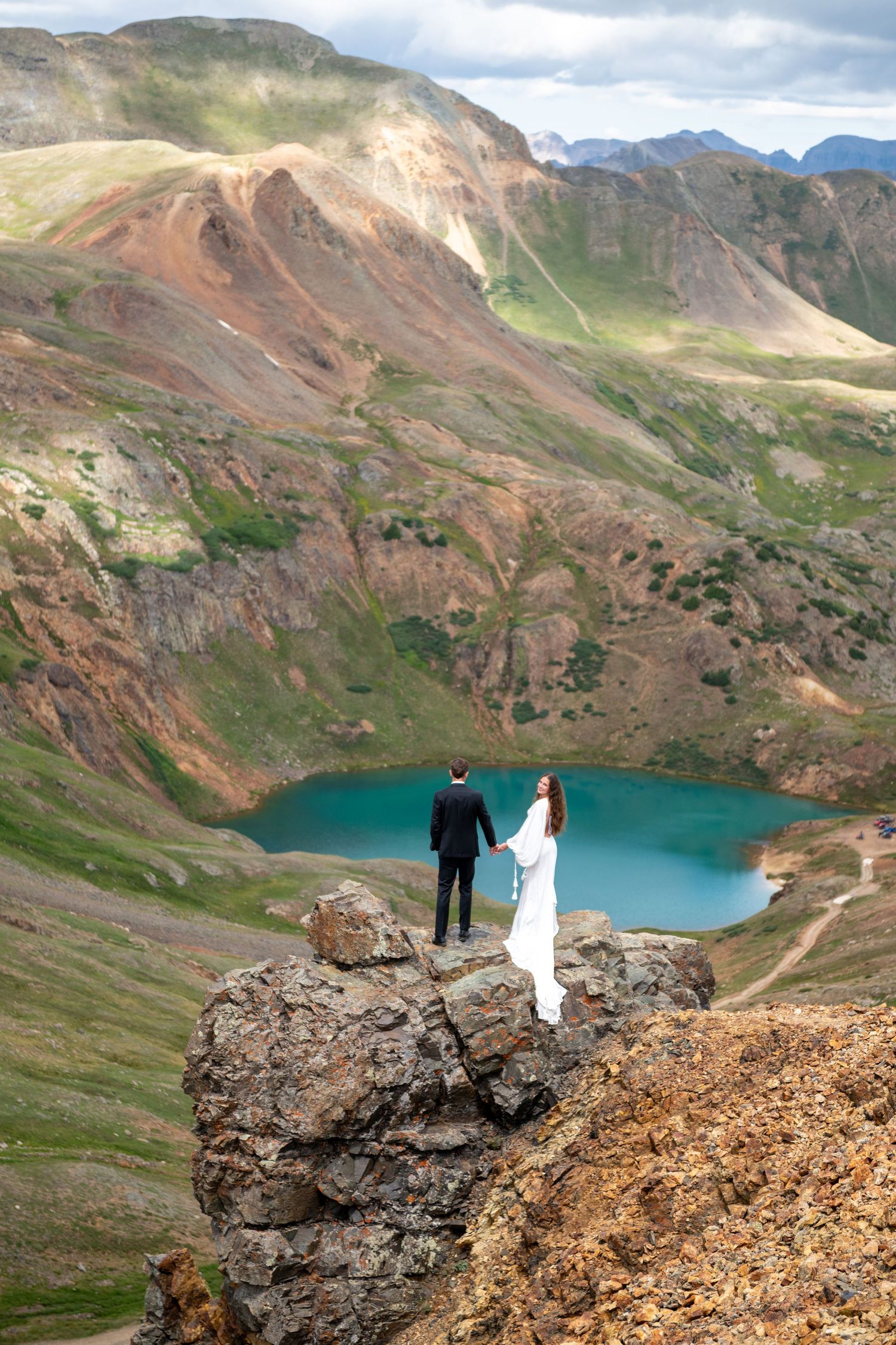 A bride and groom stand on a rocky cliff overlooking a turquoise alpine lake surrounded by colorful mountain peaks.