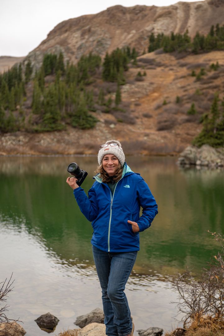 Woman in blue North Face jacket holds camera by a green mountain lake with evergreen trees and rocky hills.