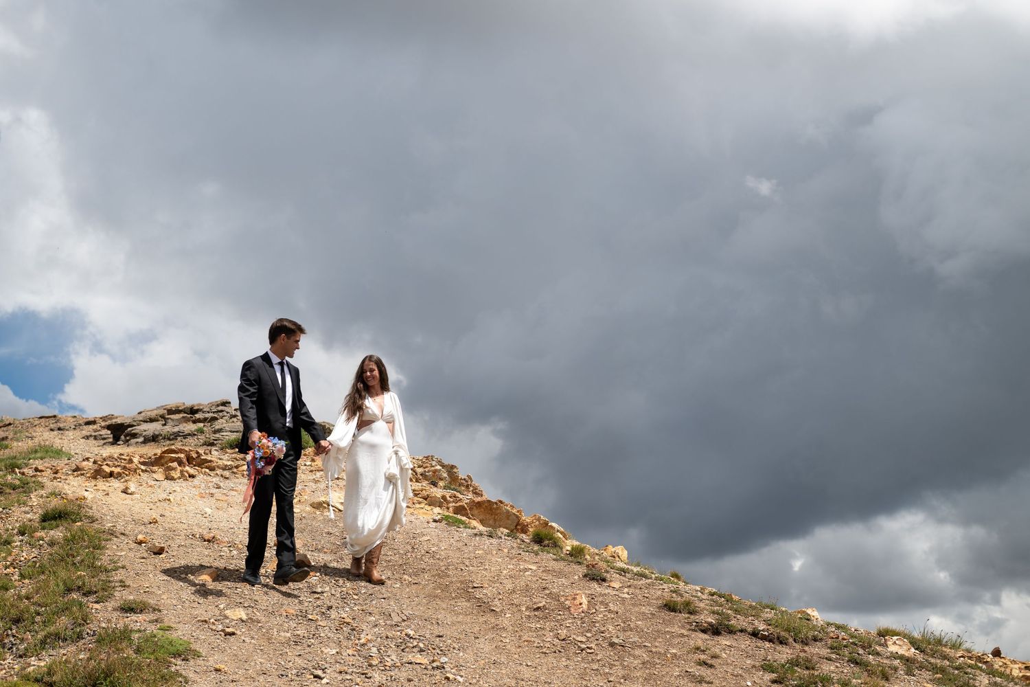 Bride and groom holding hands walking on rocky hillside under dramatic stormy clouds on their wedding day.