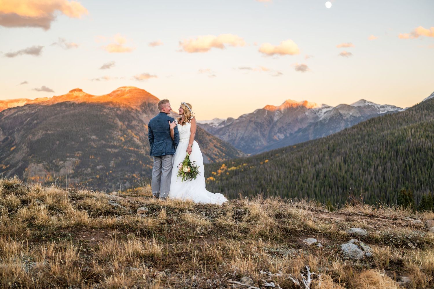 Bride and groom embrace at sunset on a mountain overlook with stunning Rocky Mountain peaks in the background.