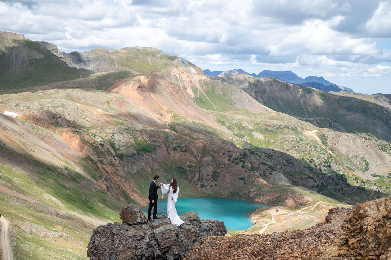 Couple in wedding attire standing on rocky mountain overlook with turquoise lake and colorful peaks backdrop.