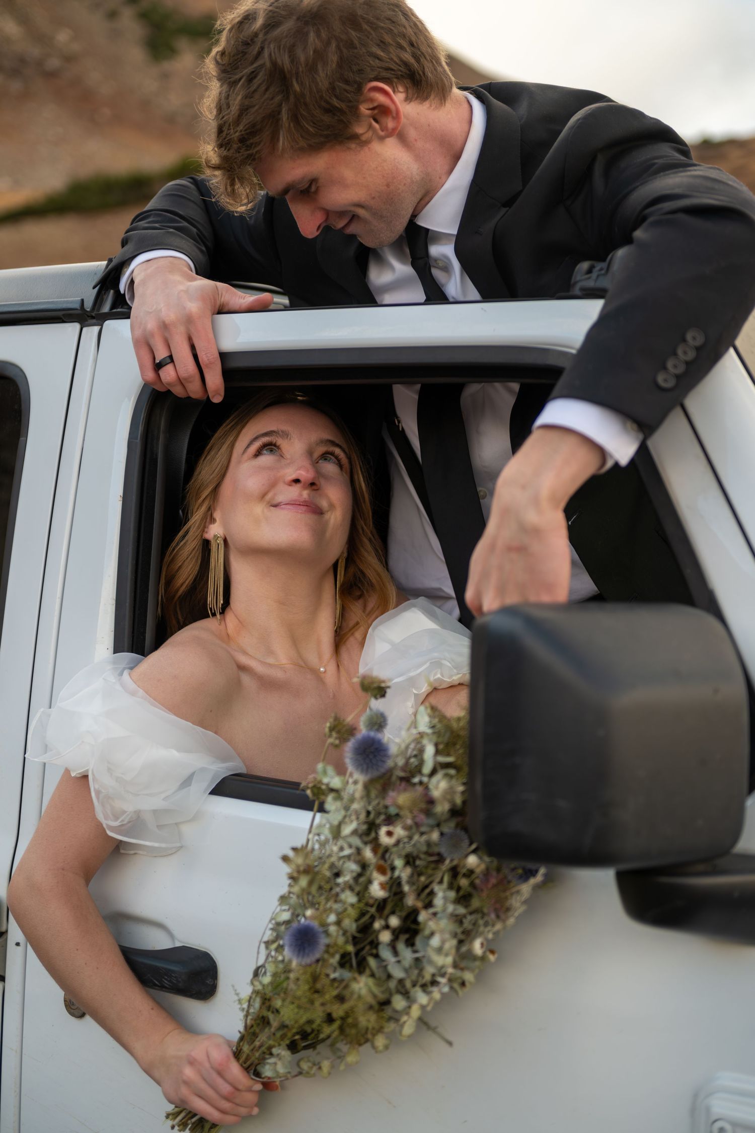 A bride leans out of a white car window holding a wildflower bouquet as the groom gazes down at her tenderly.