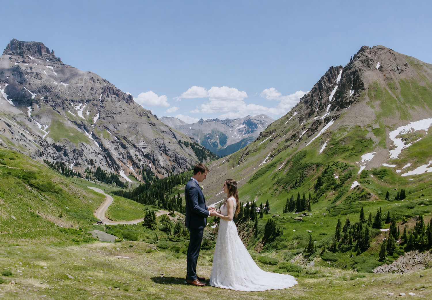 Couple exchanging vows at an outdoor mountain wedding ceremony with lush green valleys and snow-capped peaks in background.