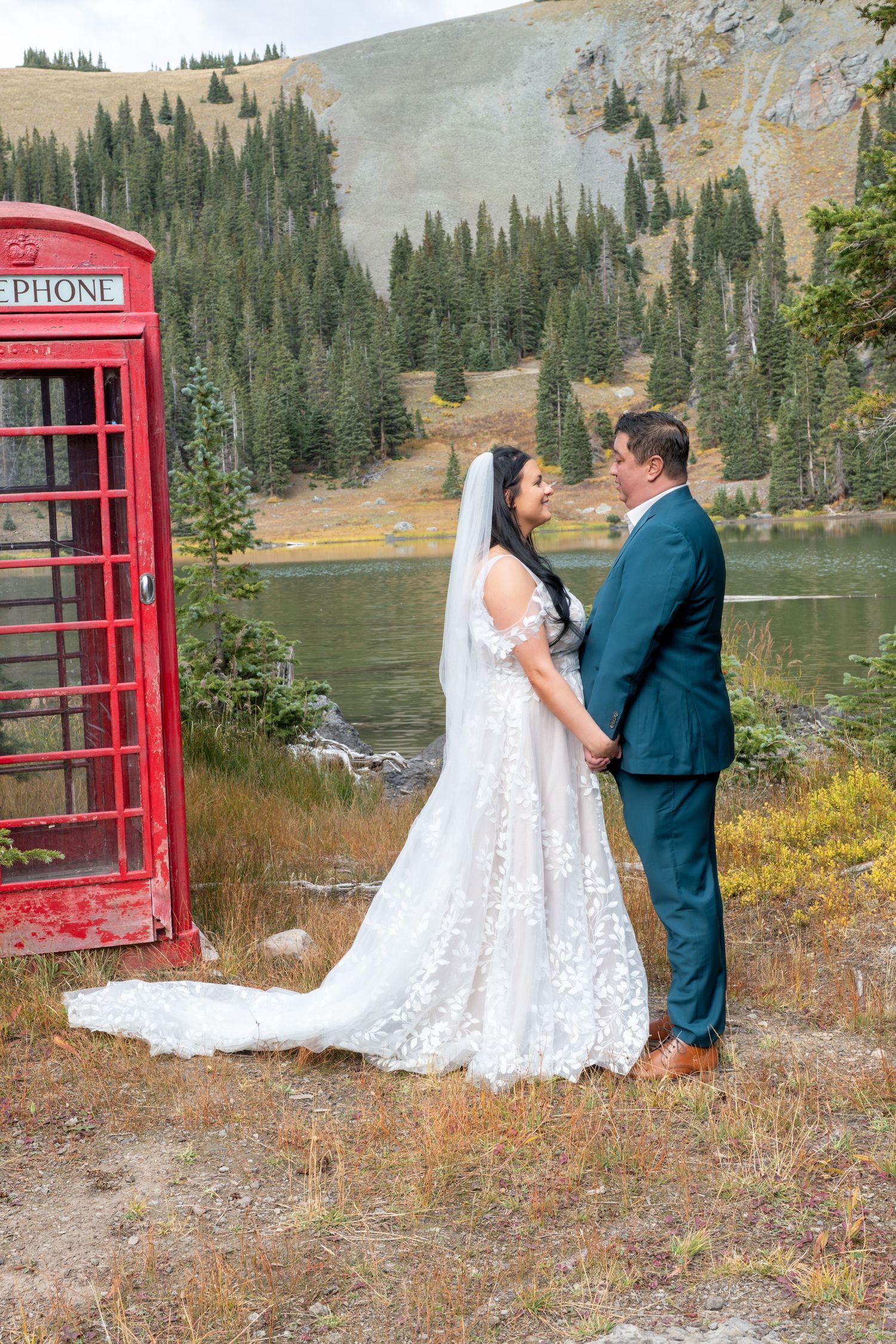 A bride and groom face each other holding hands in romantic moment by a red phone booth near a mountain lake surrounded by pine trees.