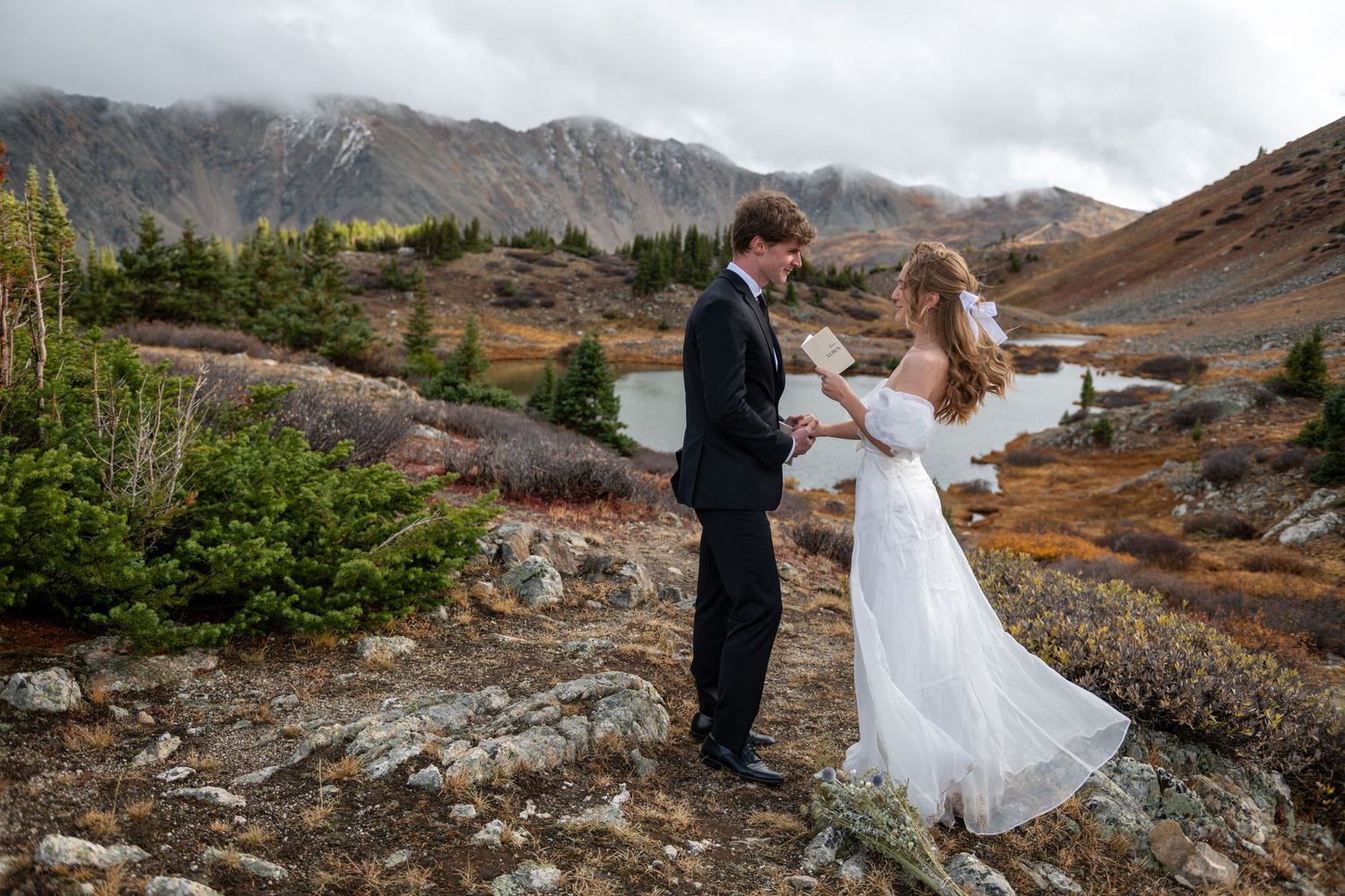 A bride reads vows to her groom during an elopement in the mountains with misty peaks and evergreens trees in the background.