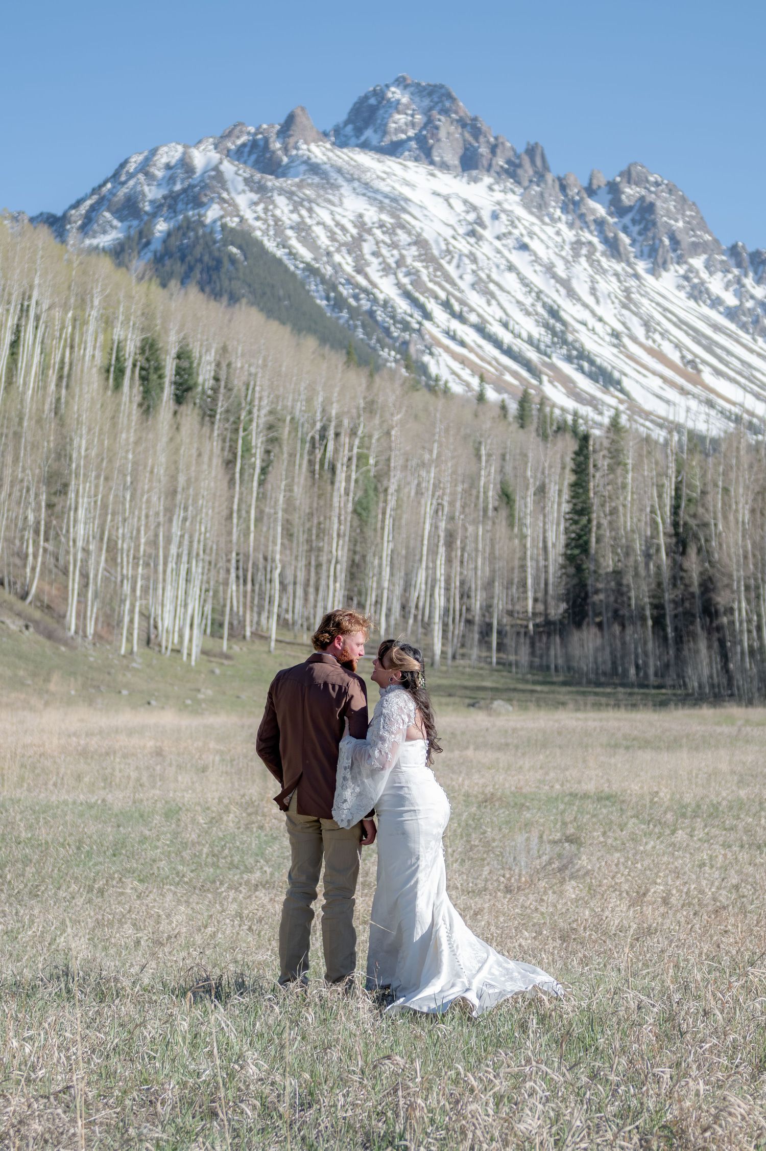 A couple shares a kiss in a mountain meadow, with snow-capped peaks and aspen trees behind them.