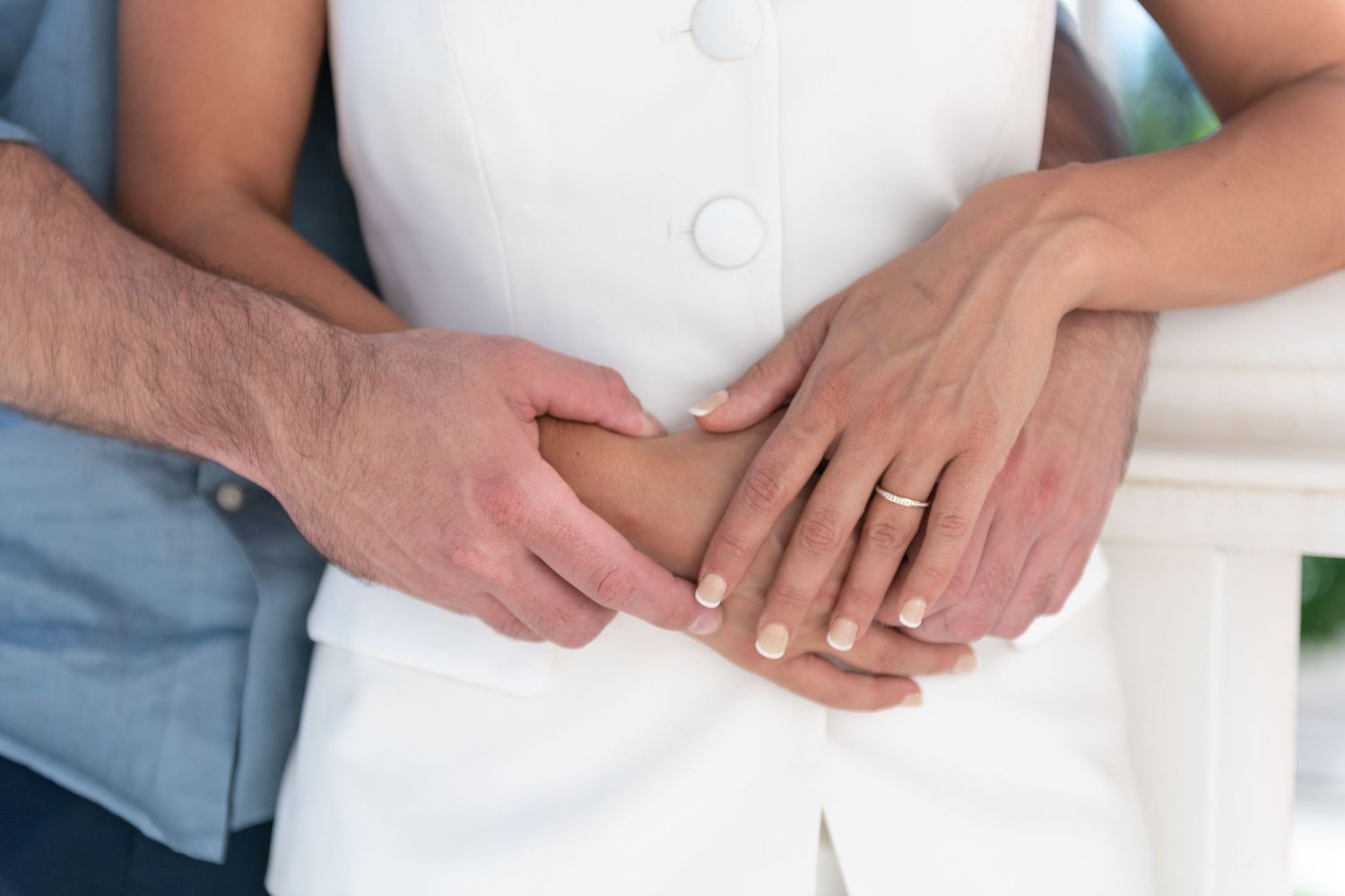 A newly wed couple embrace holding hands together, woman wearing engagement ring and white dress, romantic outdoor setting.