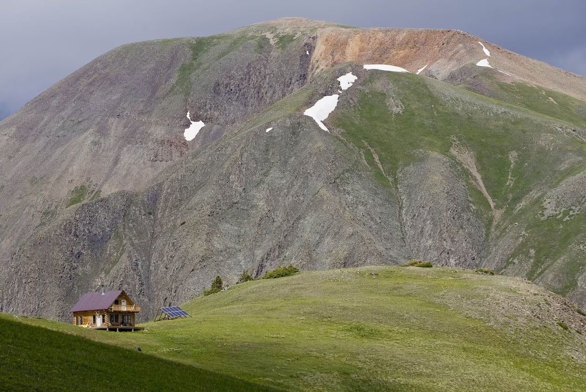 A wooden Airbnb cabin sits in an alpine meadow above treeline beneath rugged mountain peaks with patches of snow near Silverton.