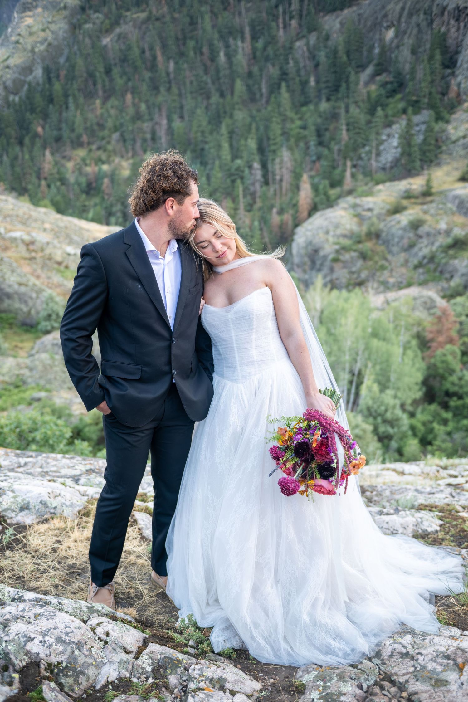 Bride in white gown holding colorful bouquet stands with groom in dark suit on rocky mountain overlook.