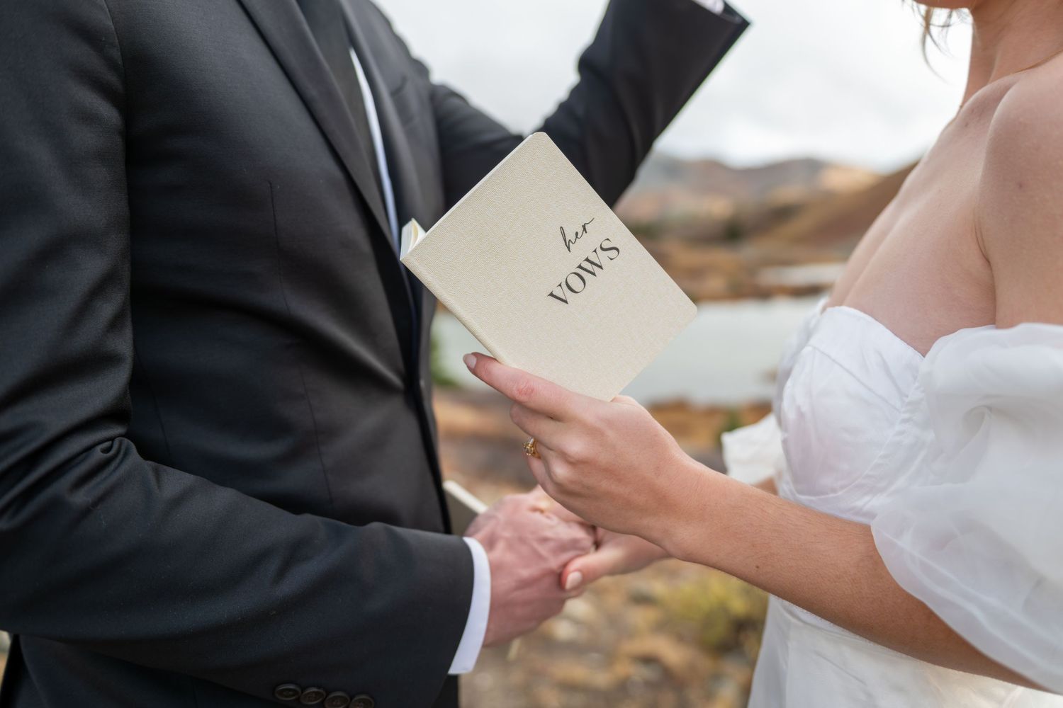 A bride holding wedding vows booklet while groom holds her hand during an outdoor elopement ceremony.