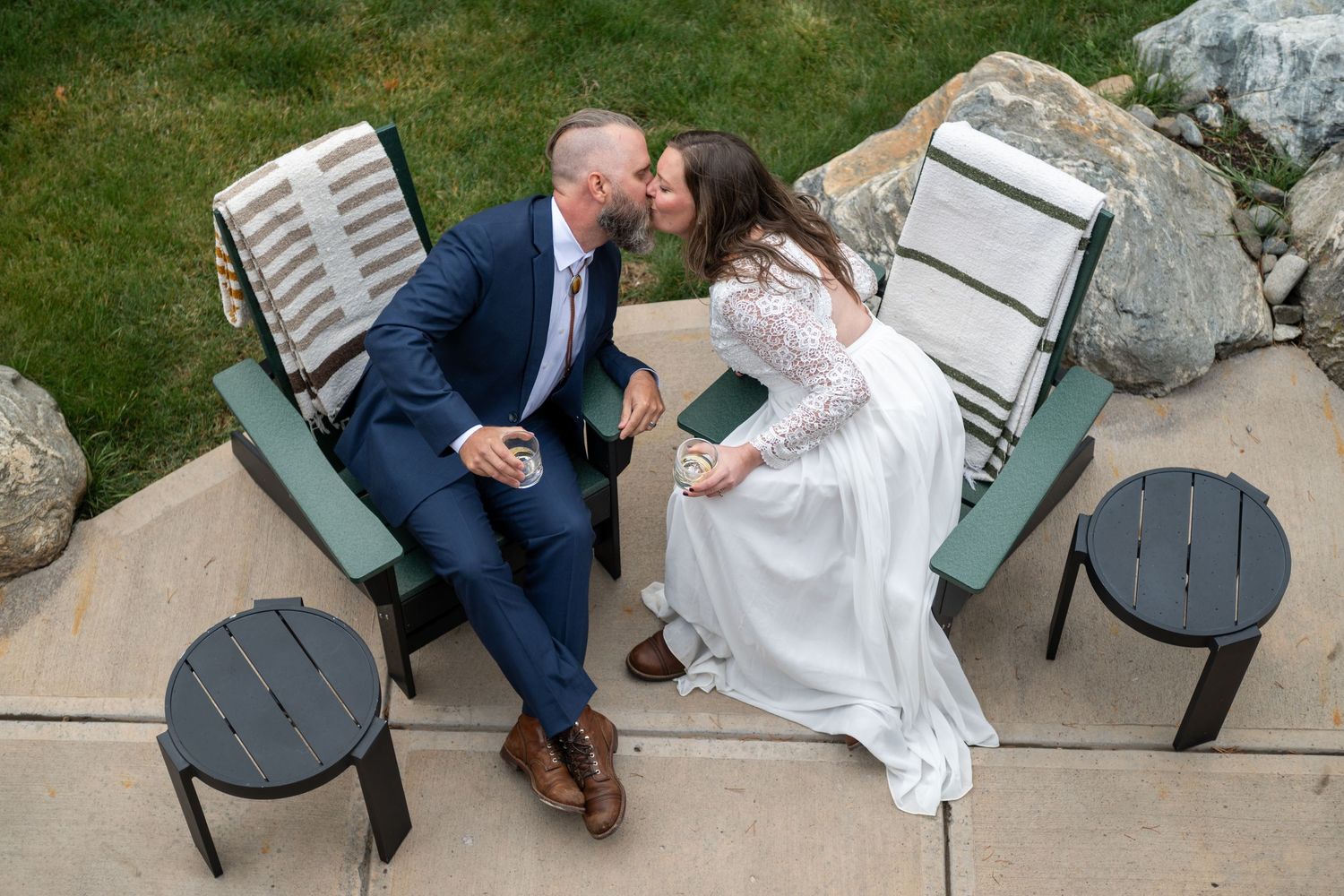 A bride and groom sharing a kiss while seated in green Adirondack chairs outdoors on their elopement wedding day.