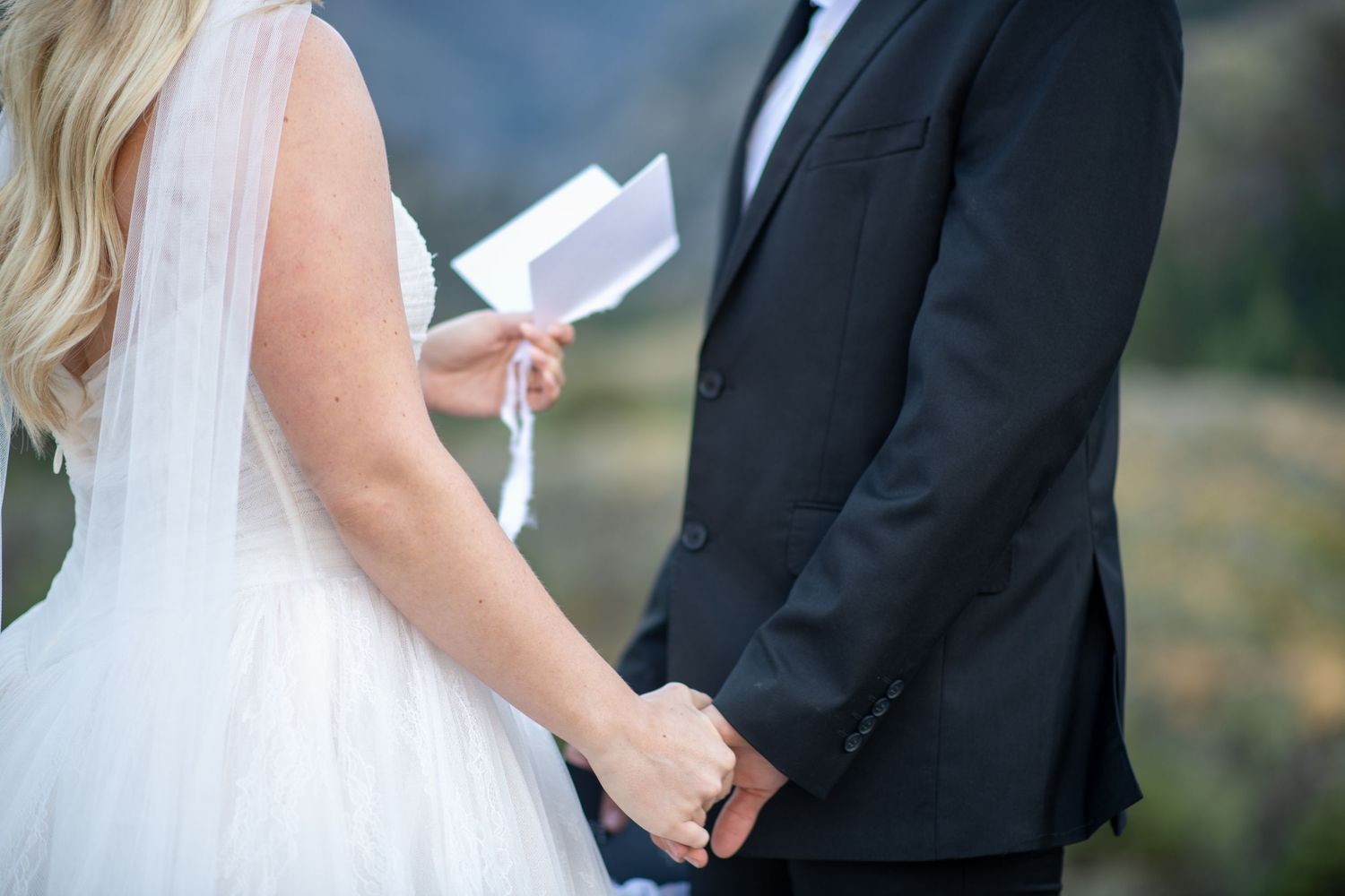 A bride and groom hold hands during outdoor wedding vows, bride in white gown holding paper vows, groom in black suit.