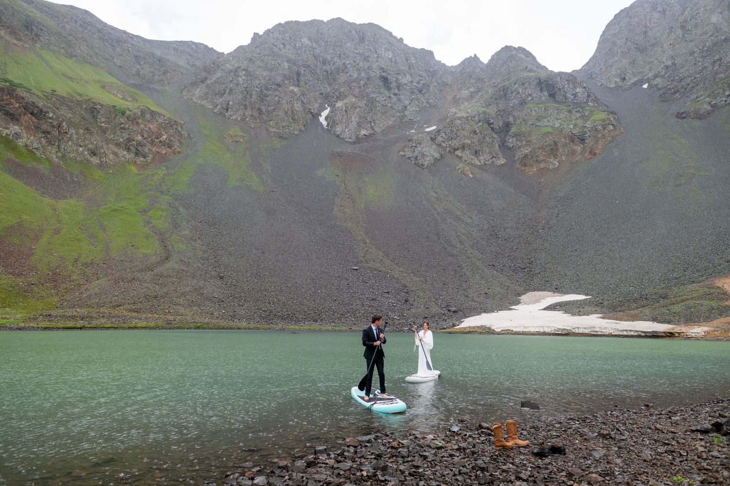 Bride and groom stand on paddleboard on alpine lake surrounded by dramatic mountain peaks.