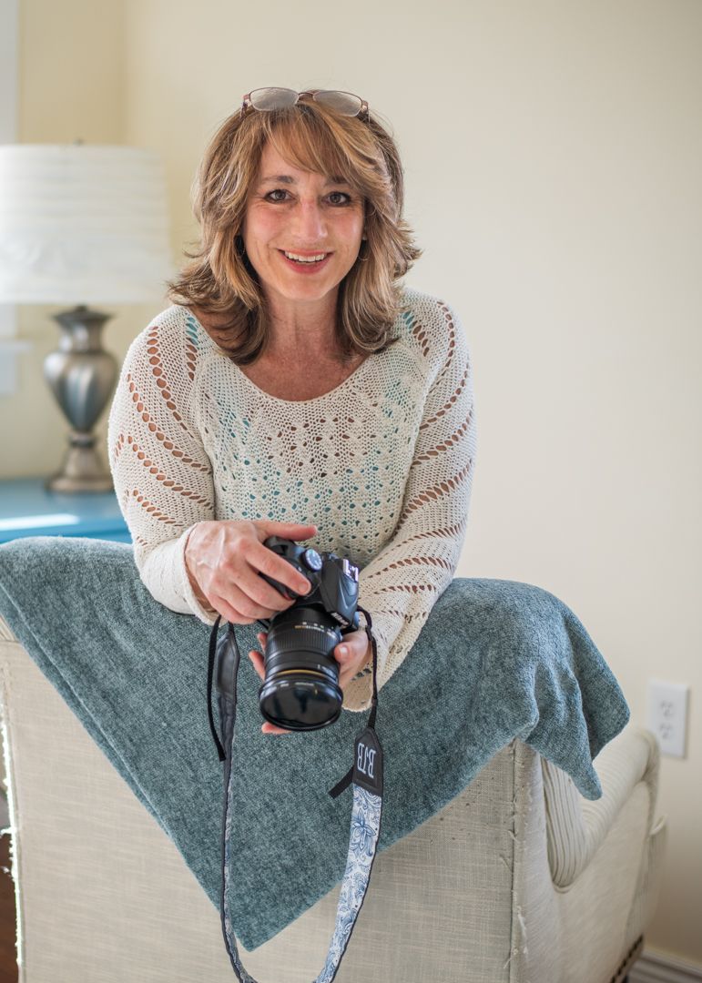 A photographer sits backward in a chair holding her camera and smiling for the photo. She is wearing a white top in a cream room.