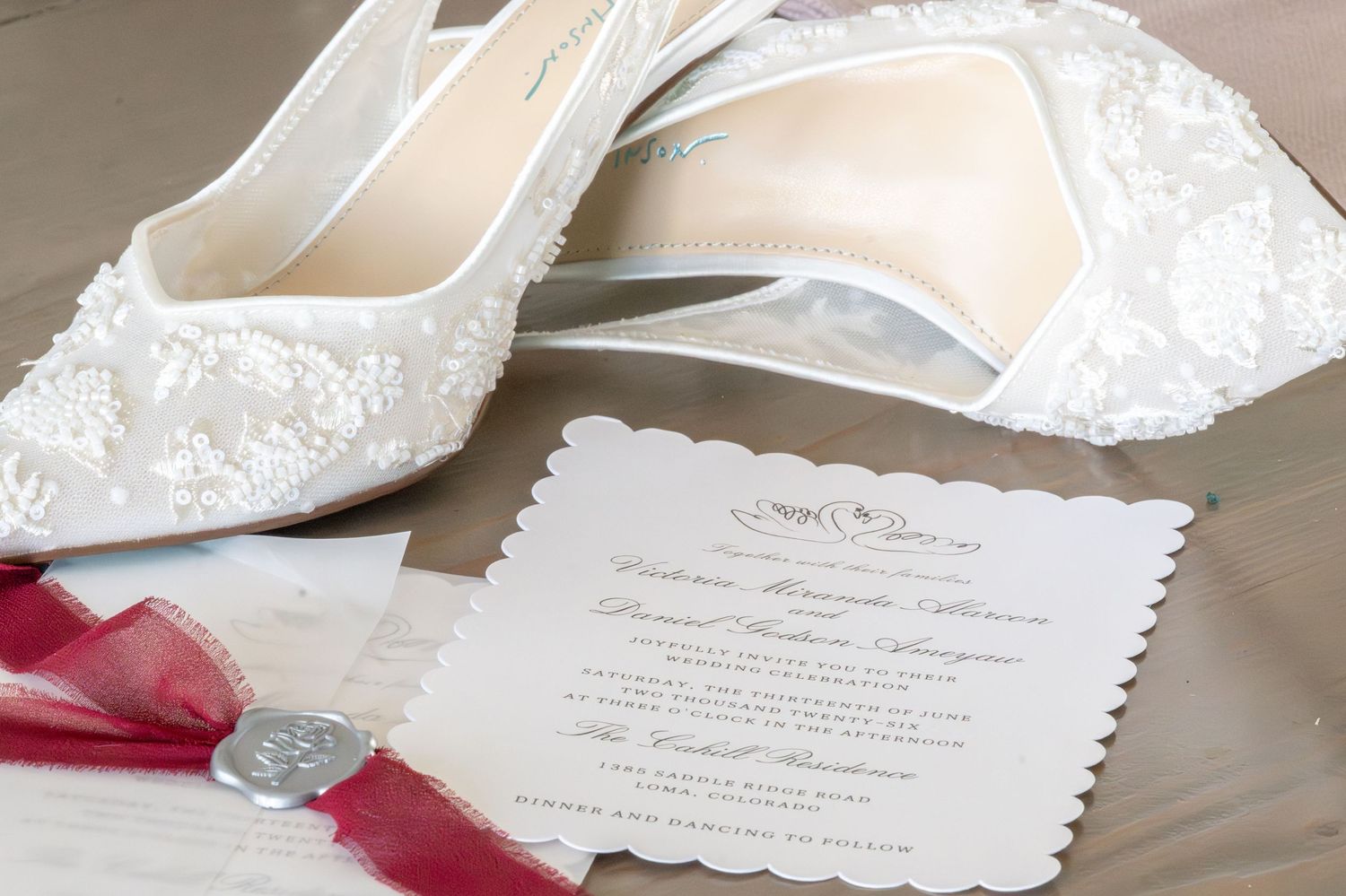 White lace bridal heels displayed beside an elegant wedding invitation with red ribbon on a soft neutral background.