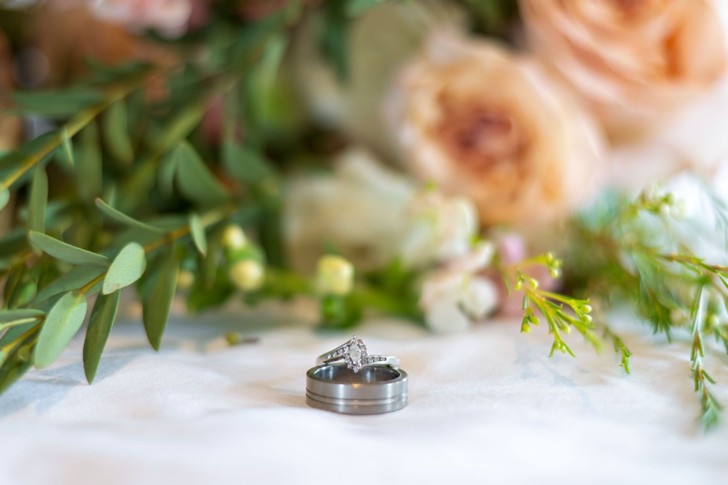 Three wedding rings displayed on white fabric surrounded by soft pink roses and green eucalyptus foliage.