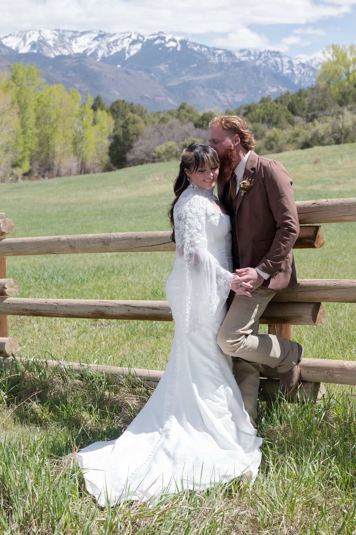 A bride and groom are nuzzled up together by a rustic wooden fence with snow-capped mountains and green fields behind them.