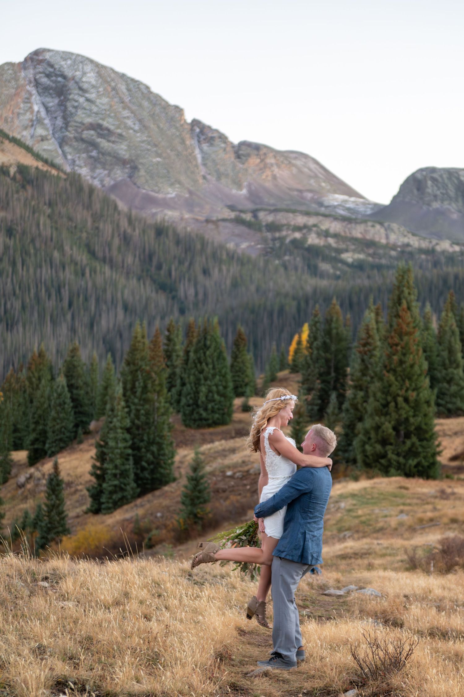 Groom lifts bride in white dress amid mountain pines and golden meadows during outdoor wedding photo session.