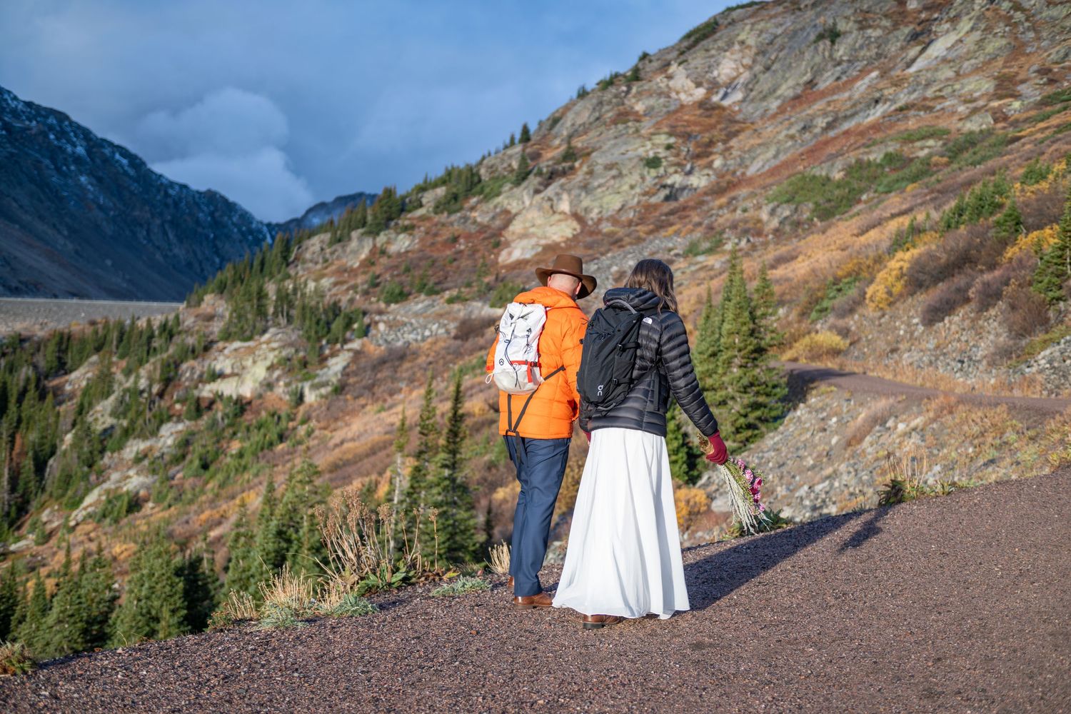 An couple walks uphill in the mountains of Colorado to say their vows by an alpine lake, They are wearing wedding attire and down coats.