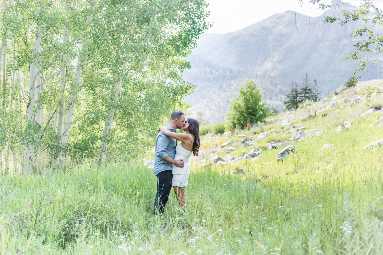 An eloping couple sharing a romantic kiss in a lush green mountain meadow surrounded by aspen trees, white flowers and scenic peaks.