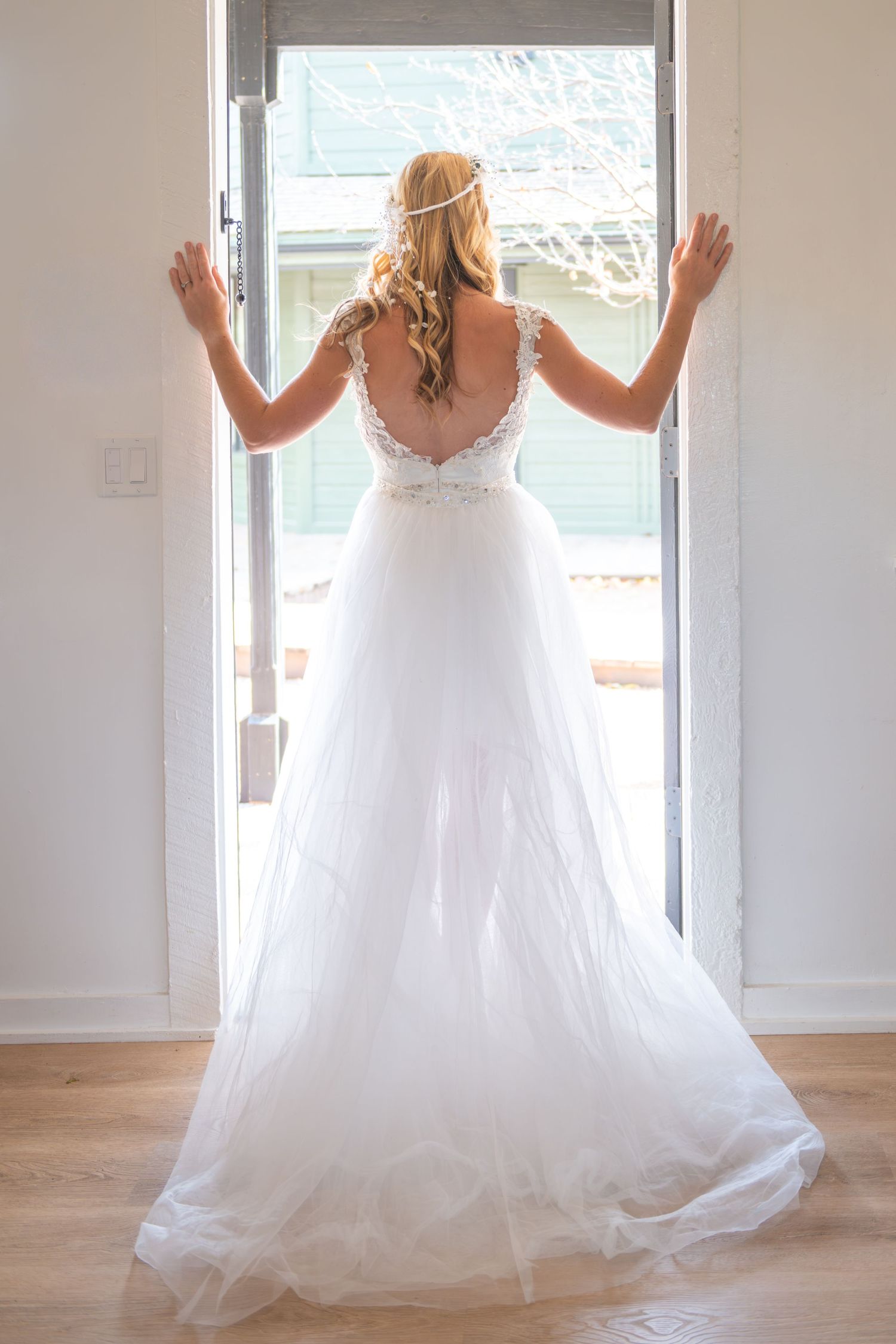 Bride in white lace wedding gown stands at open door, showing elegant backless detail and flowing tulle skirt.
